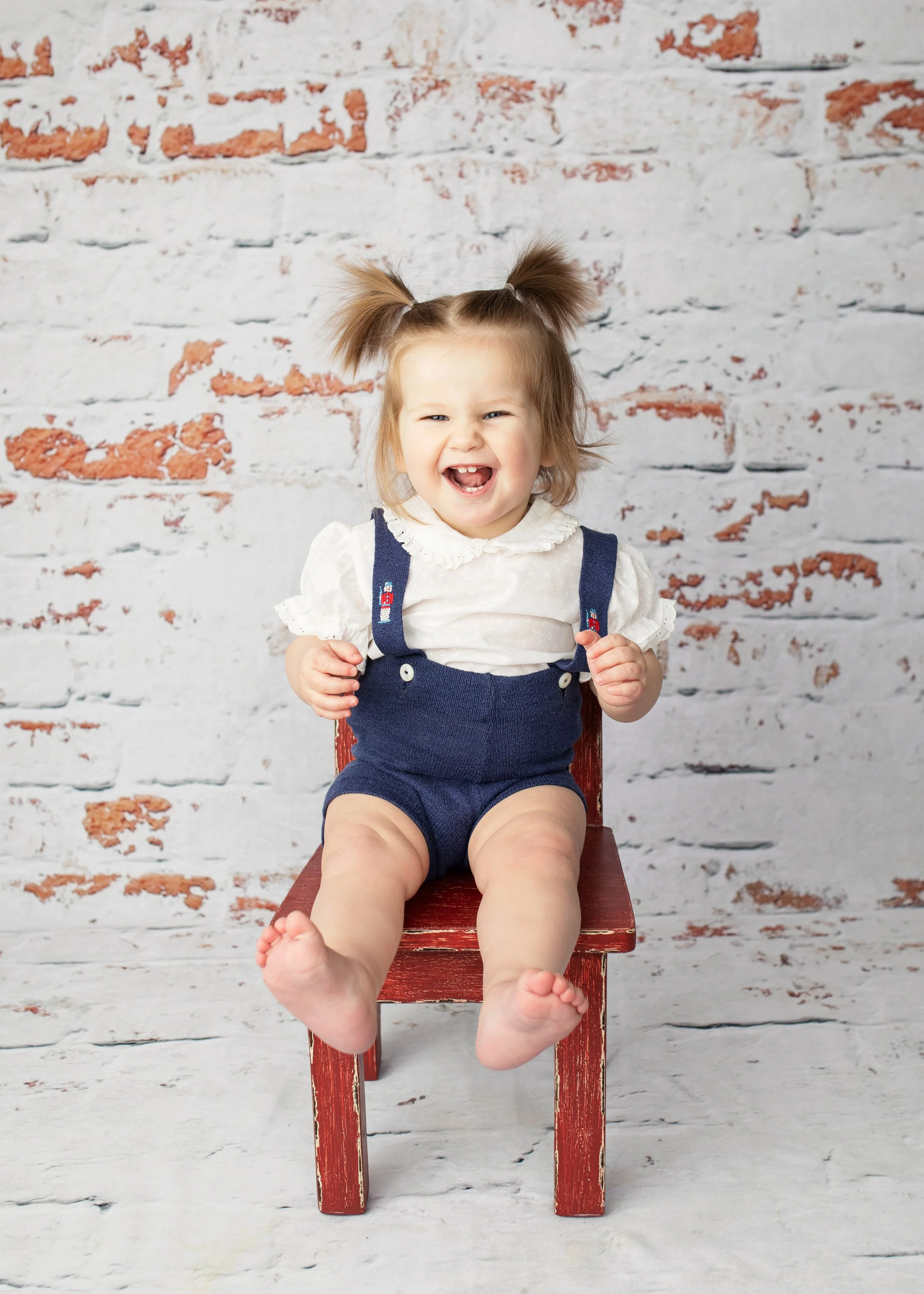 A happy young girl with pigtails, wearing a white blouse with ruffled collar and navy overalls, sitting on a small red chair in front of a white brick wall with some peeling paint.