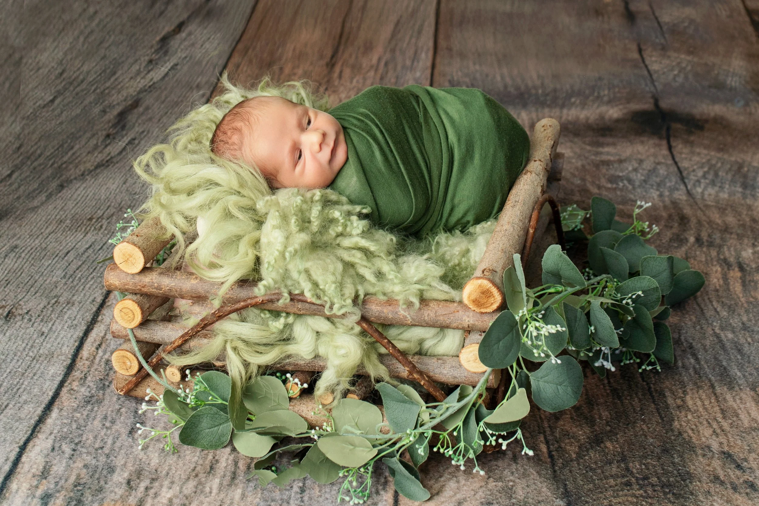 A baby with light greenish curly hair lying on a small wooden bed, wrapped in a green blanket, surrounded by green leaves on a wooden floor.