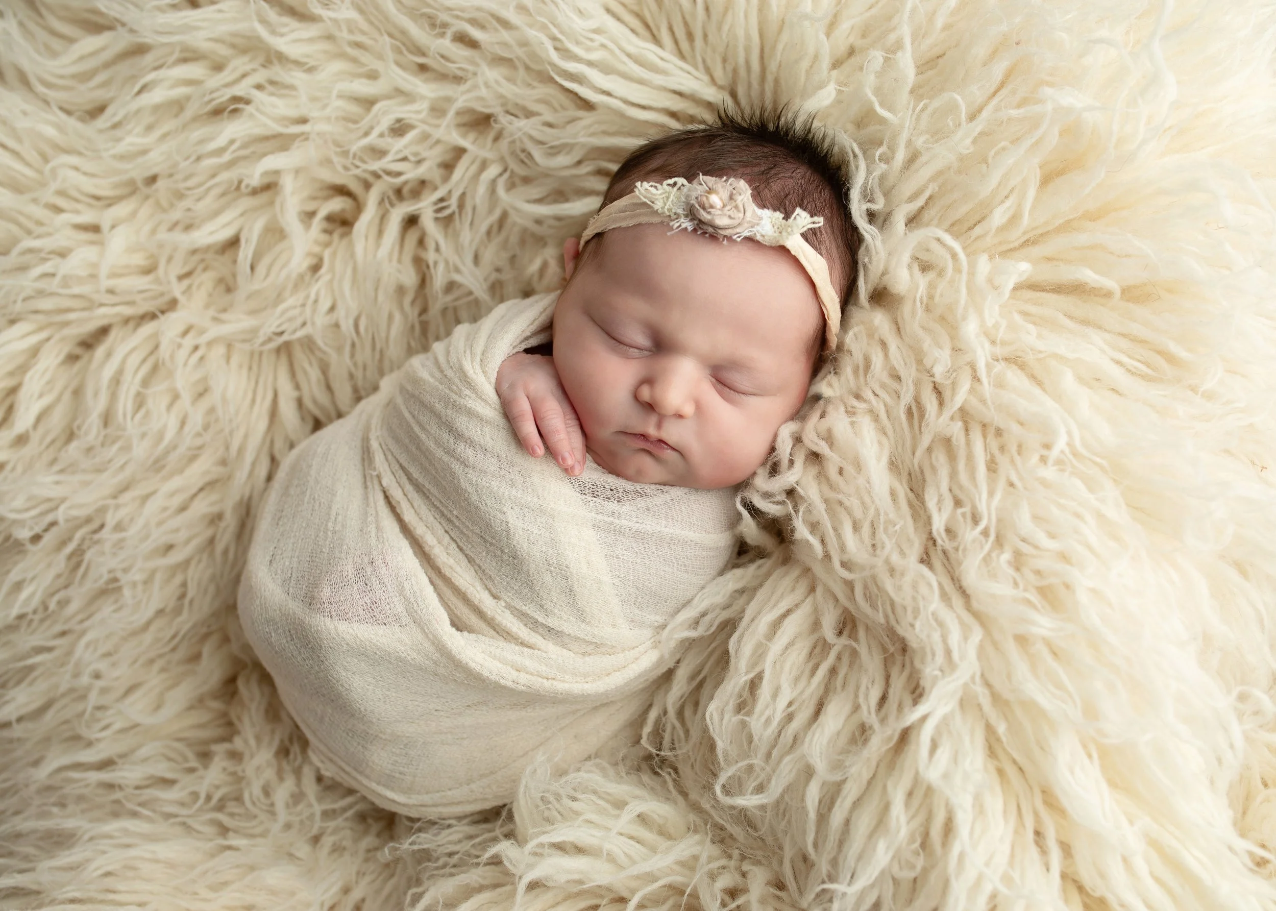 Close-up of a sleeping newborn baby lying on a fluffy, cream-colored blanket, wearing a cream-colored outfit and a headband with a small flower detail.
