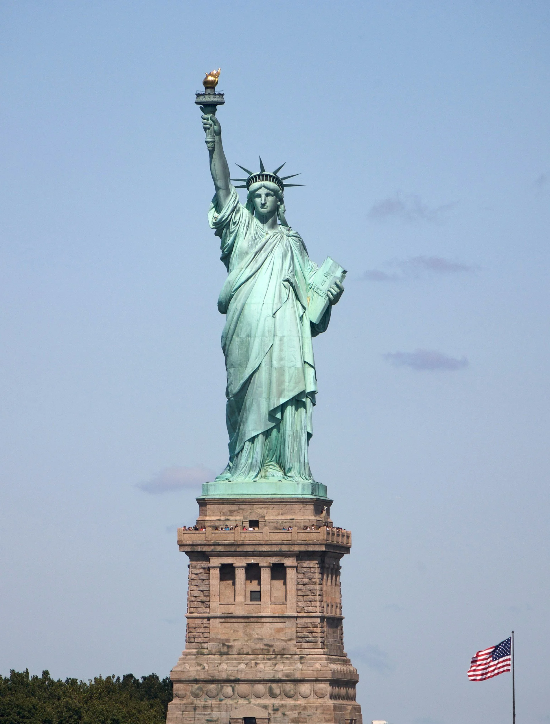 The Statue of Liberty standing on a stone pedestal, holding a torch in her right hand and a book in her left, with an American flag flying nearby against a blue sky with some clouds.
