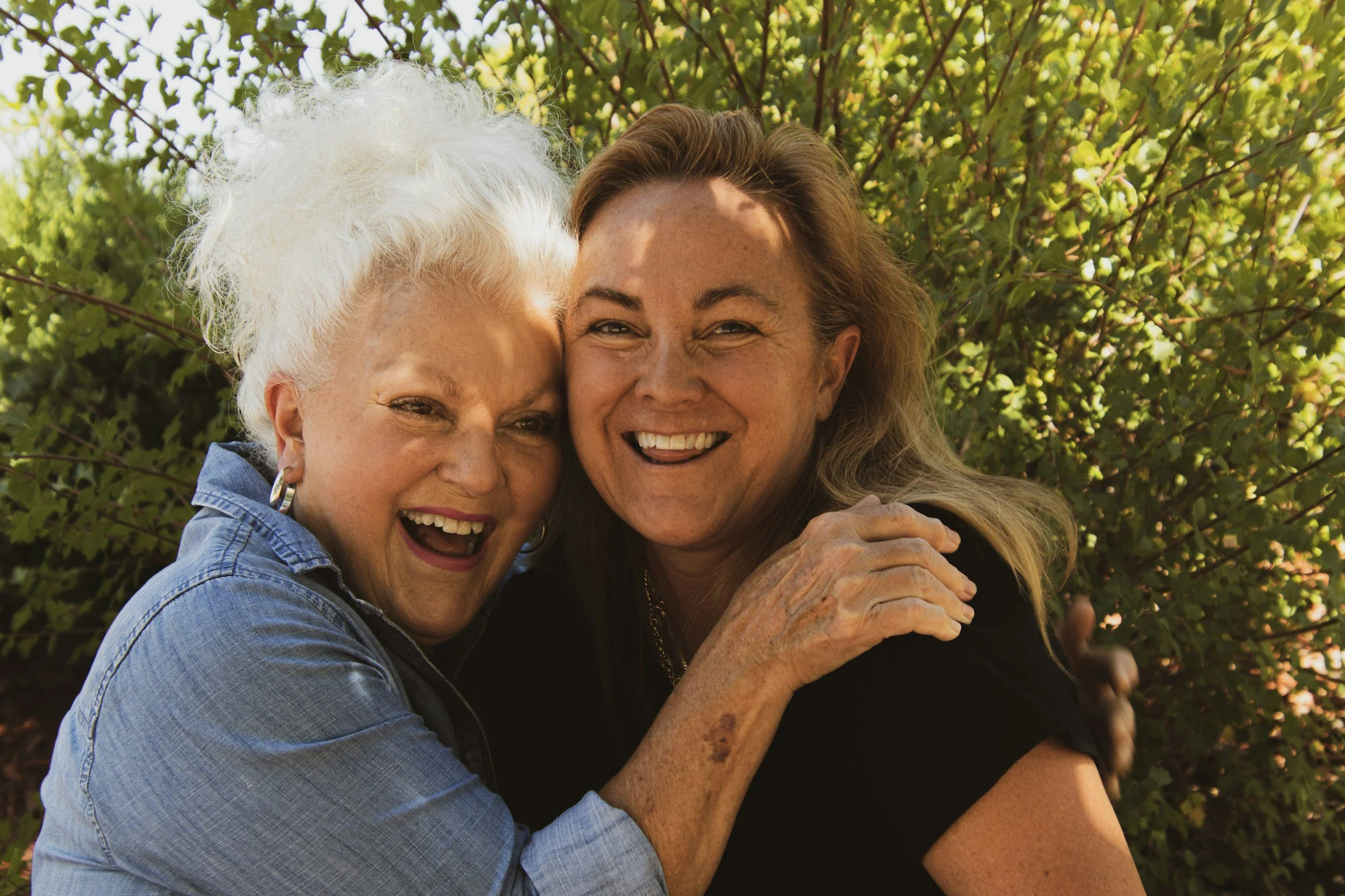 Two women, one older with white hair and one younger with brown hair, smiling and embracing outdoors with green leaves in the background.