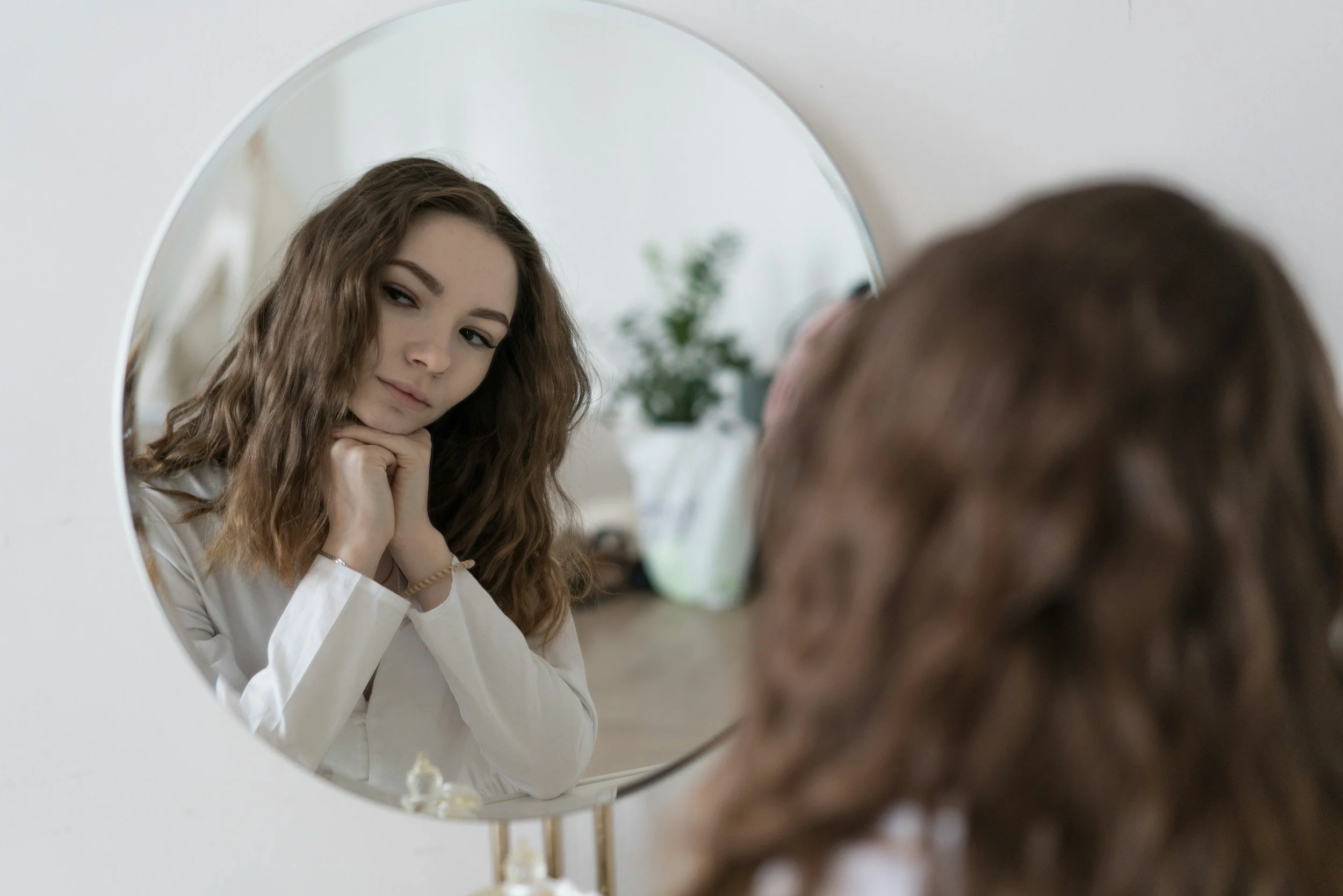 Young woman with wavy brown hair looking at herself in a round mirror, resting her chin on her hand, in a bright room with white walls and a plant in the background.