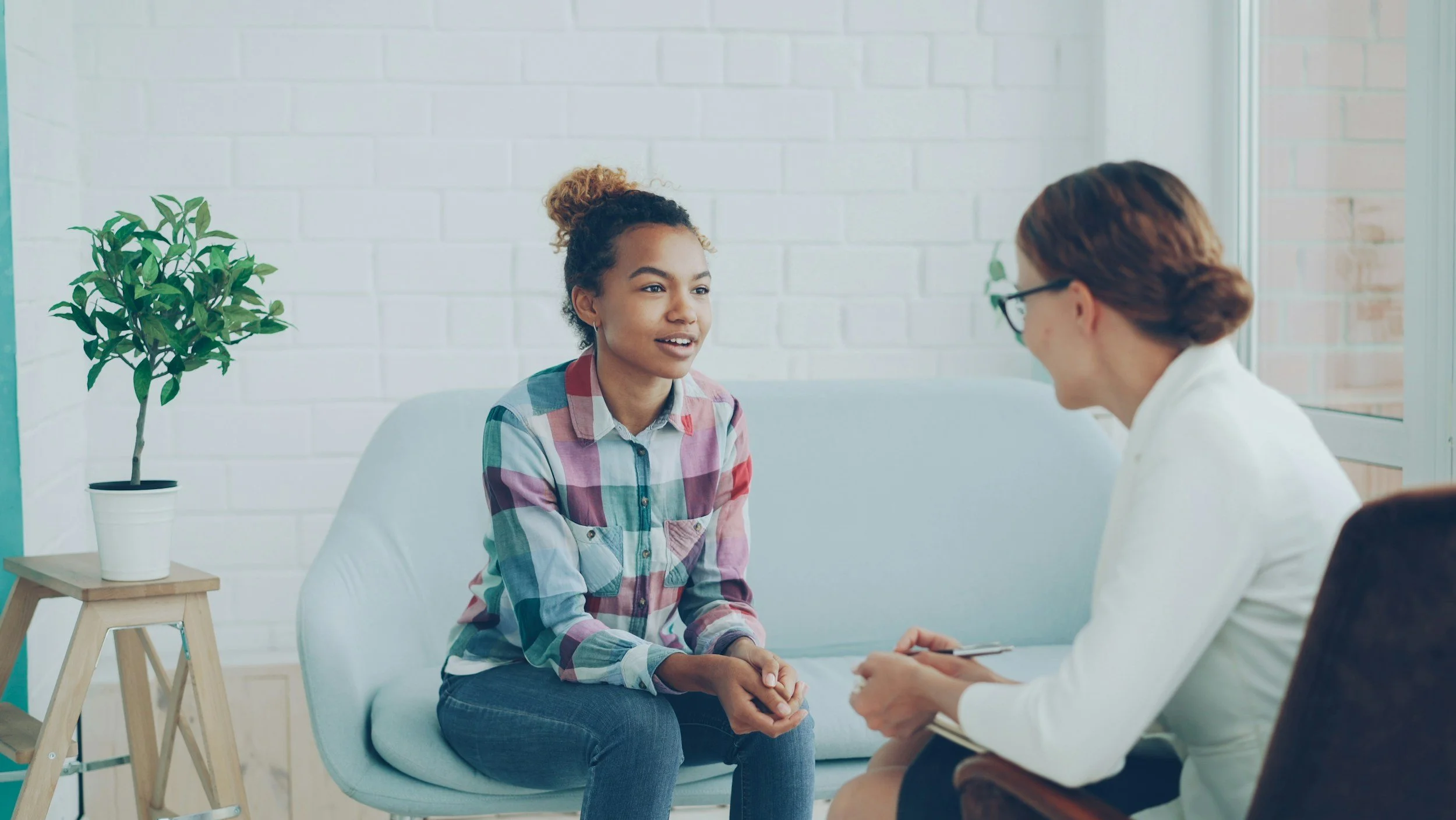 A young woman sitting on a light blue couch, talking with a healthcare professional or counselor, in a bright room with white brick walls and a potted plant on a small wooden side table.