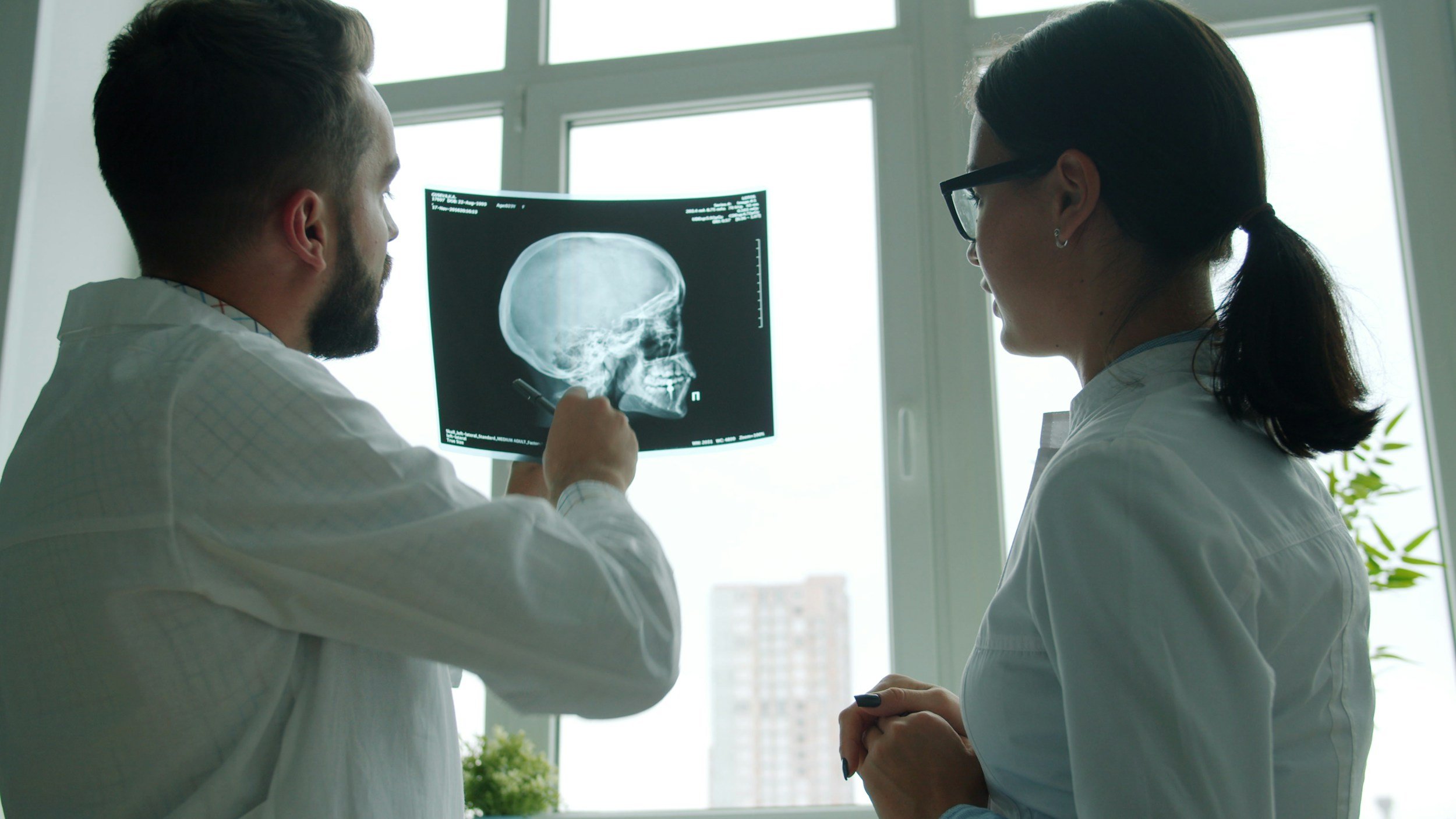 Medical professionals examining an X-ray of a human skull in a well-lit room with large windows.