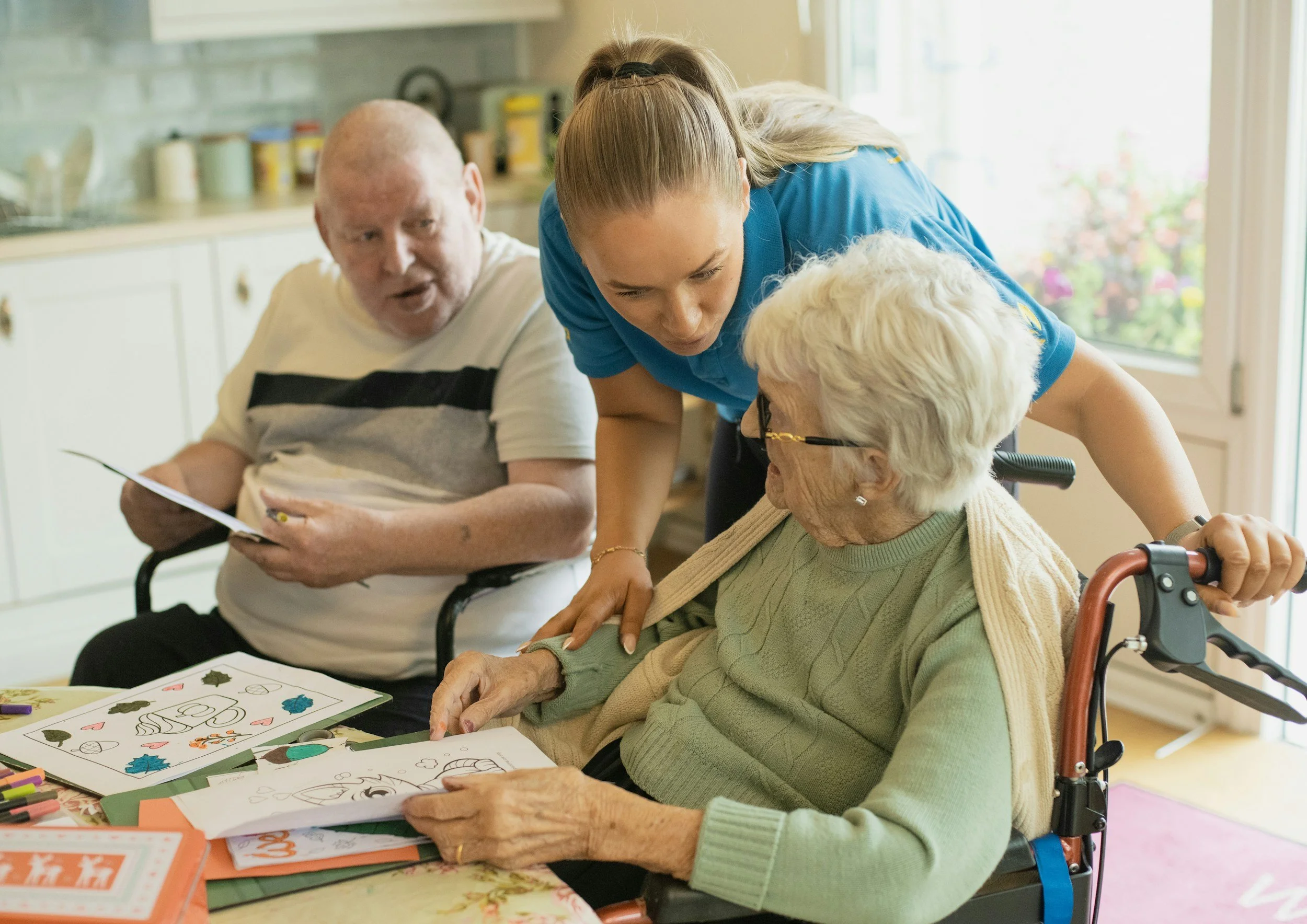 An elderly woman with white hair and glasses sitting in a wheelchair, looking at her granddaughter who is leaning over her, both engaged in a coloring activity at a table. The granddaughter is wearing a bright blue shirt and appears excited or happy. There's another person in the background, holding a paper, with a kitchen and a window with flowers outside visible in the background.