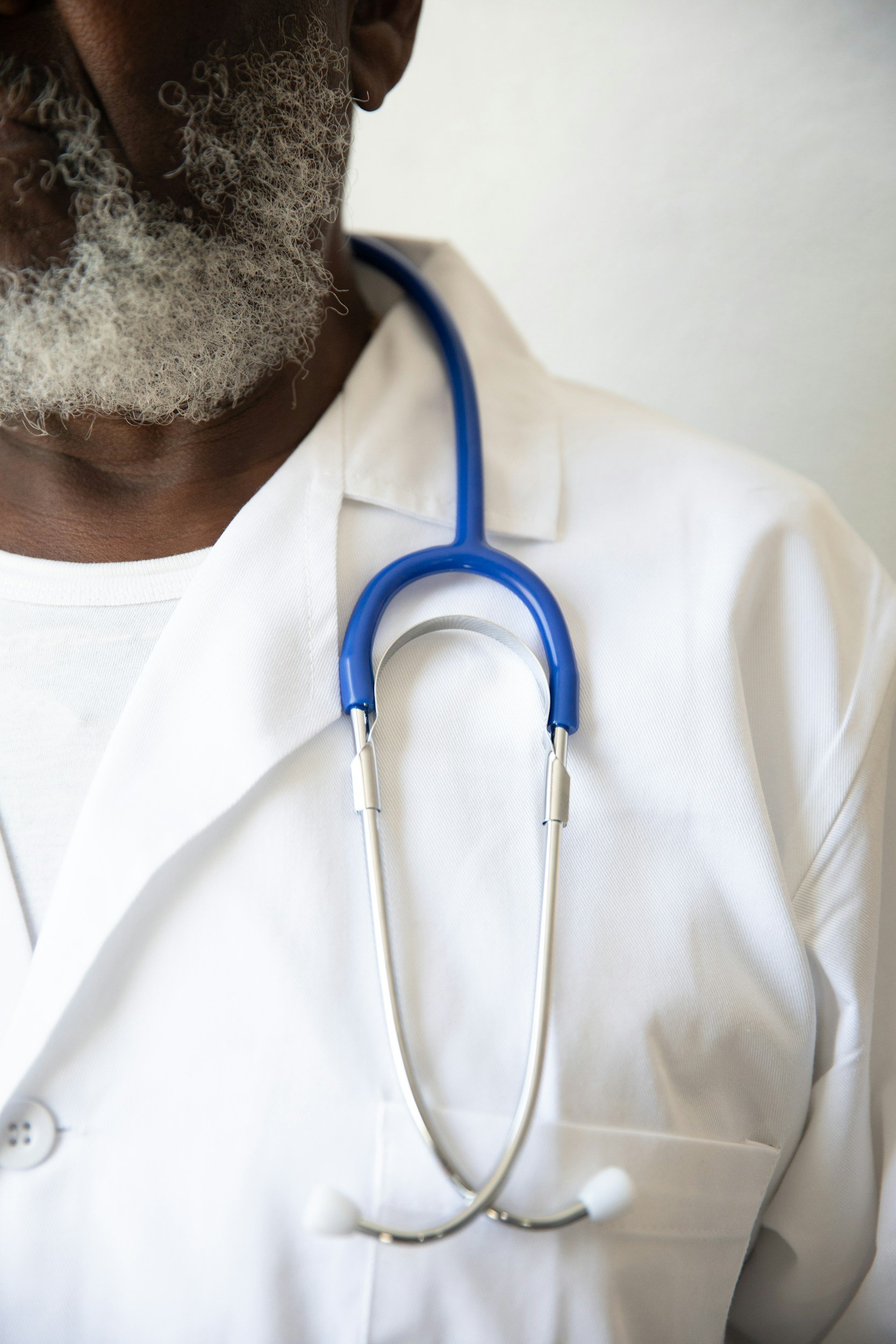 Close-up of a male doctor with a gray beard wearing a white lab coat and a blue stethoscope around his neck.