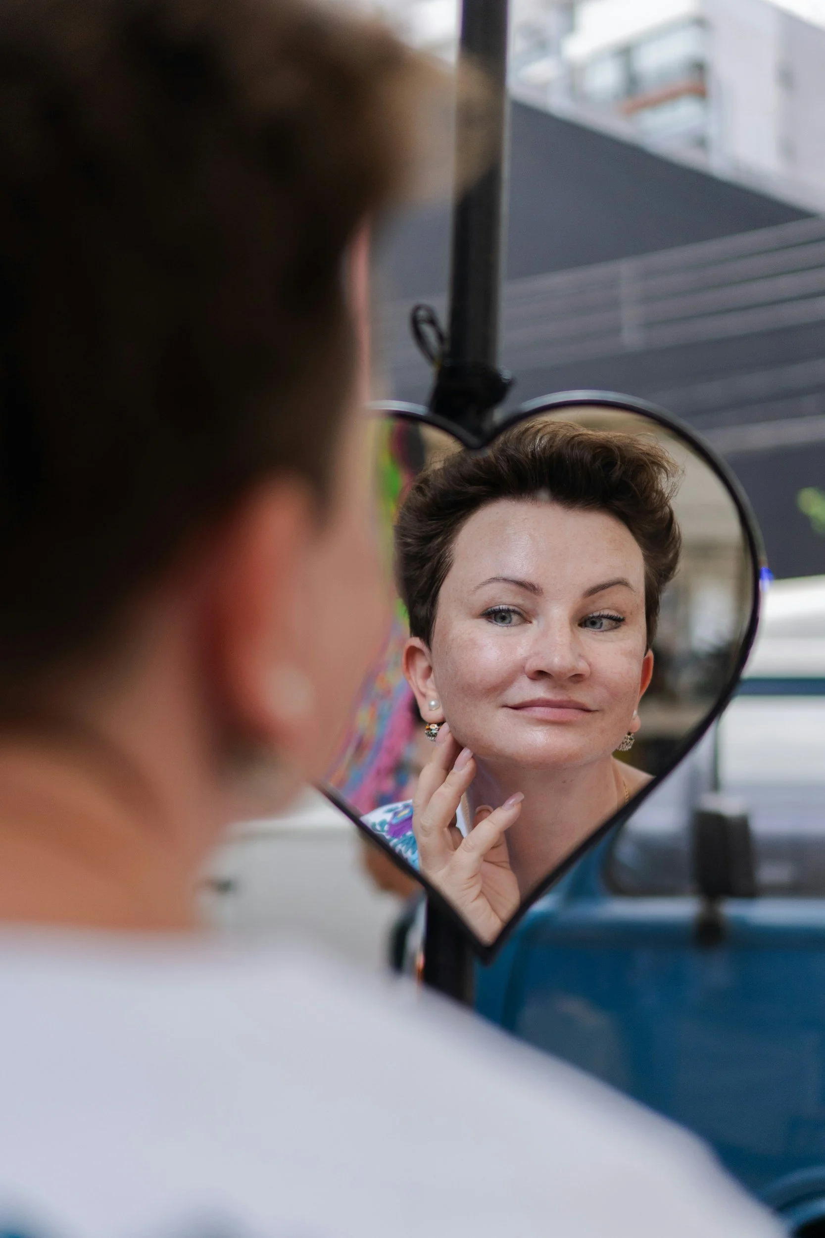 A woman with short brown hair and earrings is looking into a round mirror, touching her chin, and smiling softly. The reflection shows her face and part of her neck. The background is blurred with buildings visible outside.