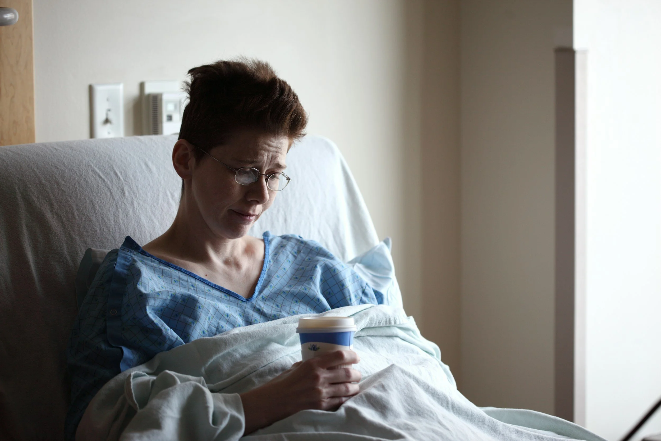 An elderly woman with short brown hair and glasses sitting in a hospital bed, holding a coffee cup, with a neutral expression, in a hospital room with beige walls and medical equipment in the background.