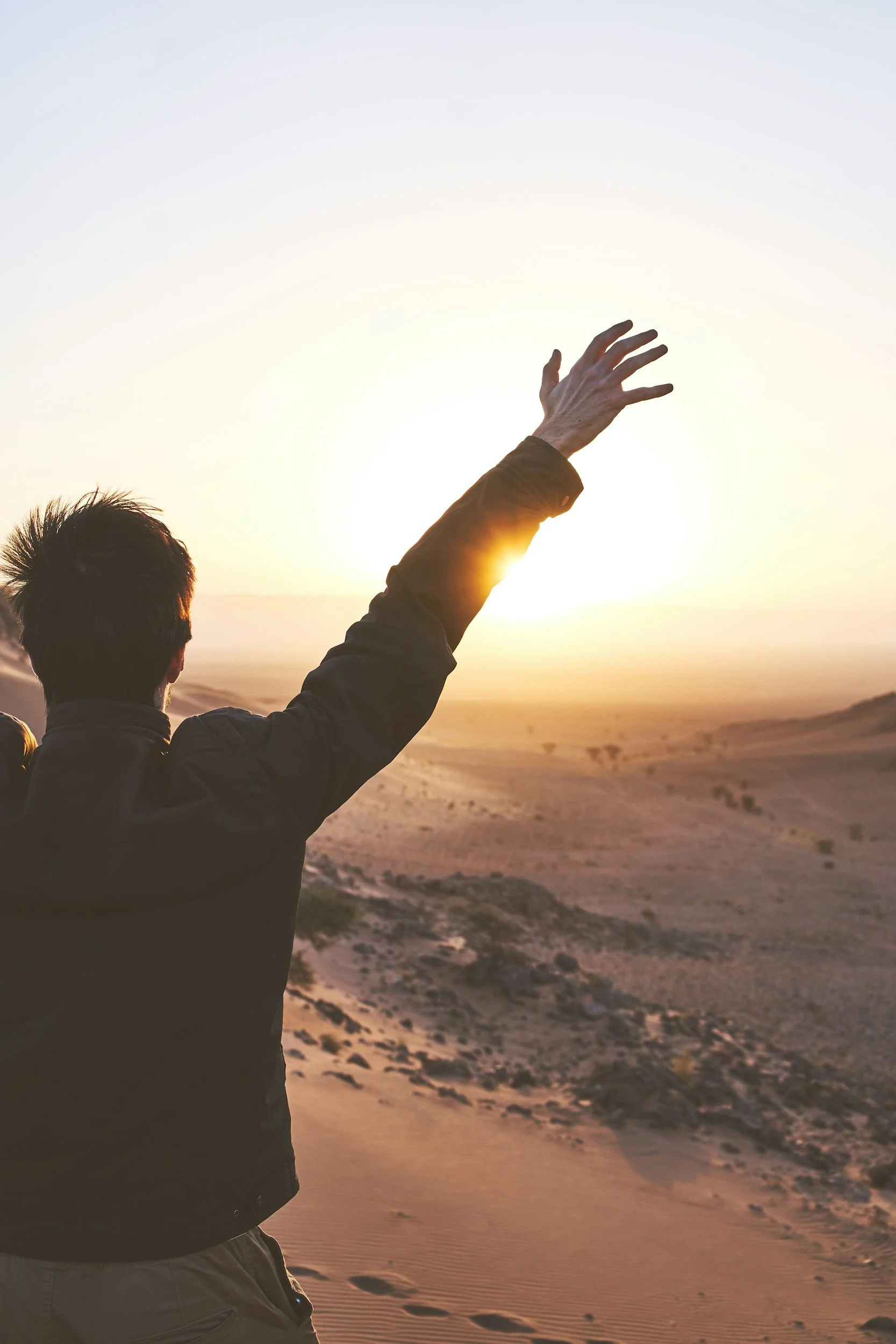 A person with short dark hair wearing a jacket, standing on a sandy desert at sunset, raising their hand towards the sky.