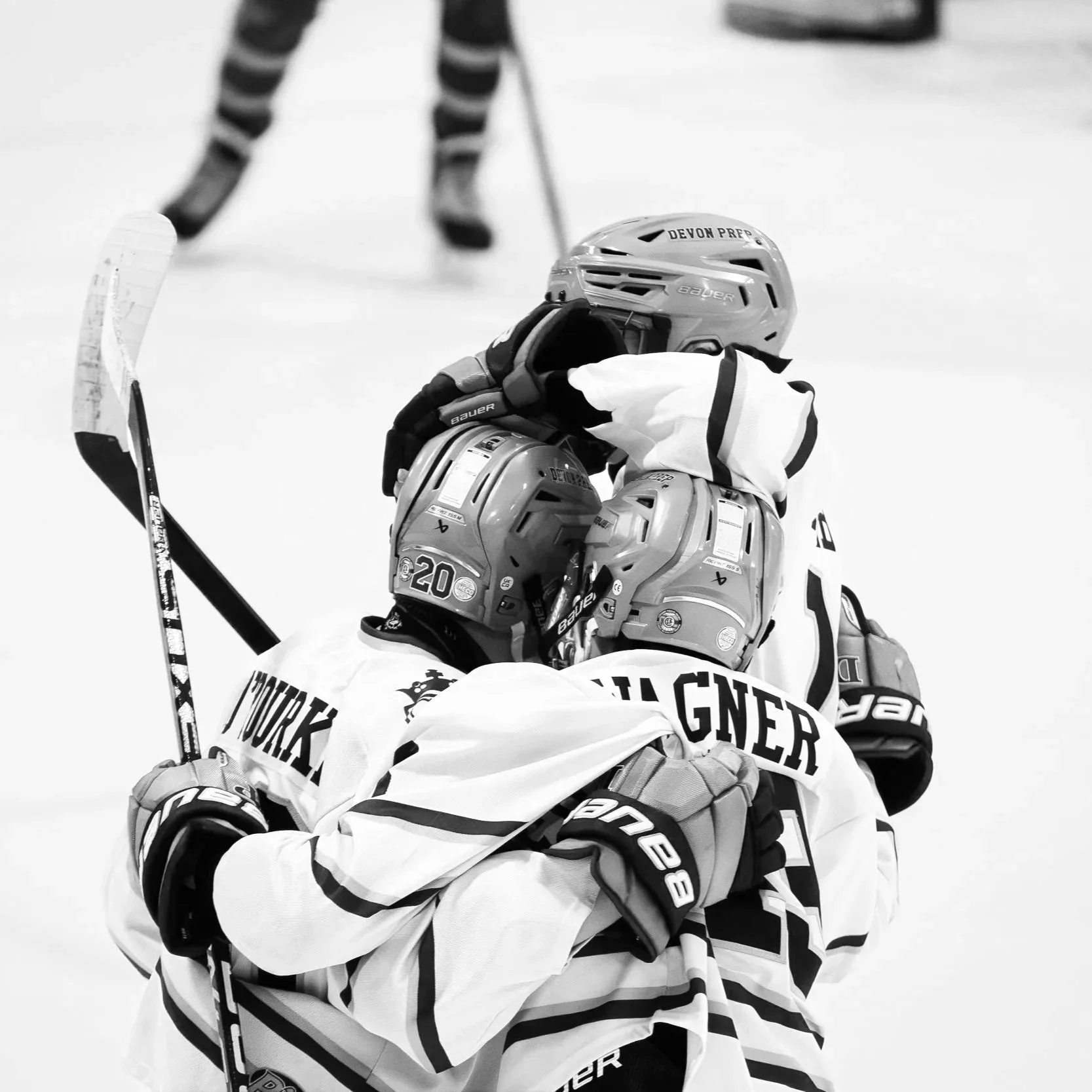 An ice hockey team celebrates on the ice with a player clutching the shoulder of a teammate, all wearing helmets and jerseys with "DEVON PREP" and "GNER" visible, with one player holding a hockey stick.