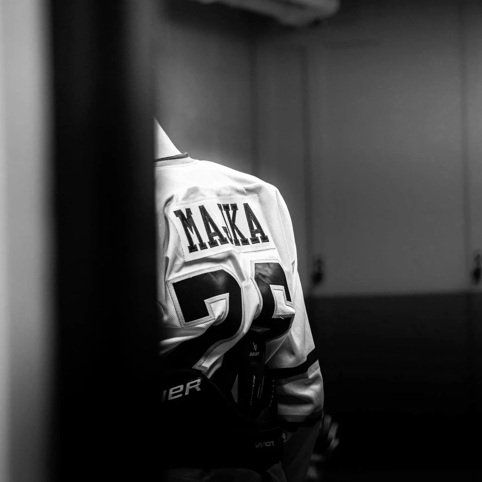 Black and white photo of a hockey player inside a locker room, partially visible from behind a door or wall, wearing a jersey with Cyrillic characters, a hockey glove, and holding a hockey stick.