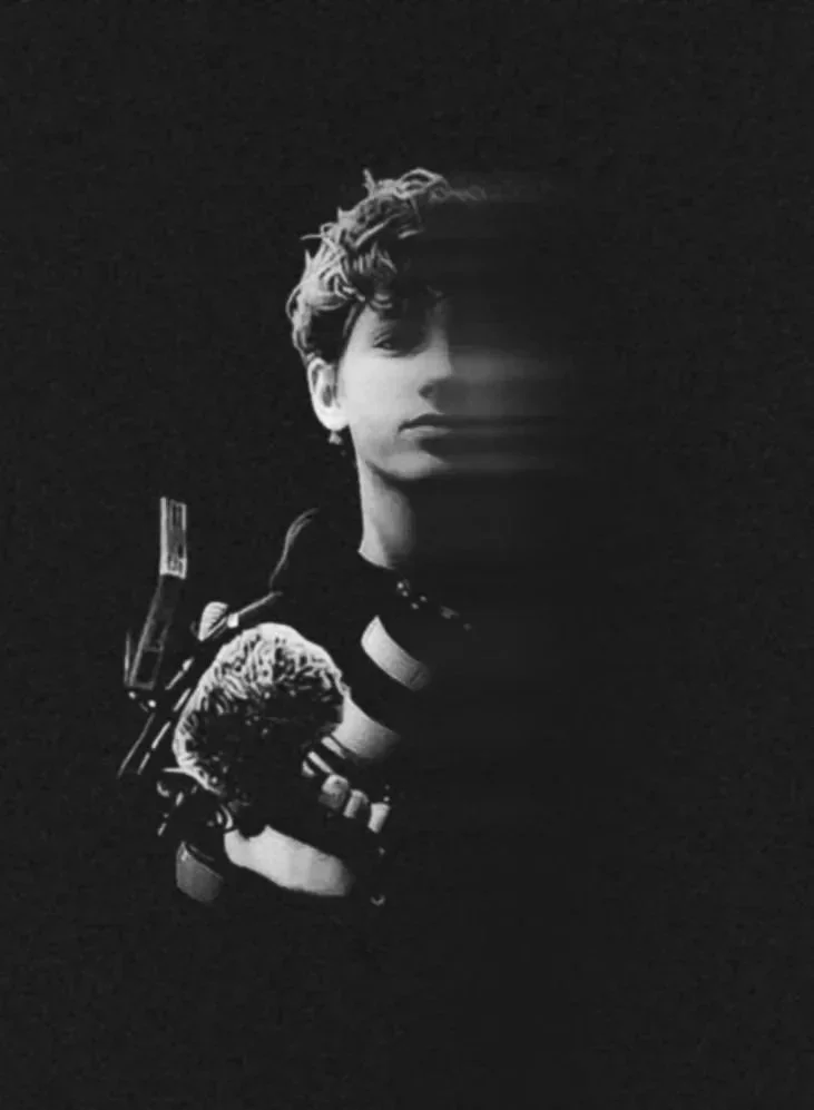 Black and white photo of a person with short, wavy hair; part of their face is shadowed and they are holding a gun with a furry object attached to it in one arm.