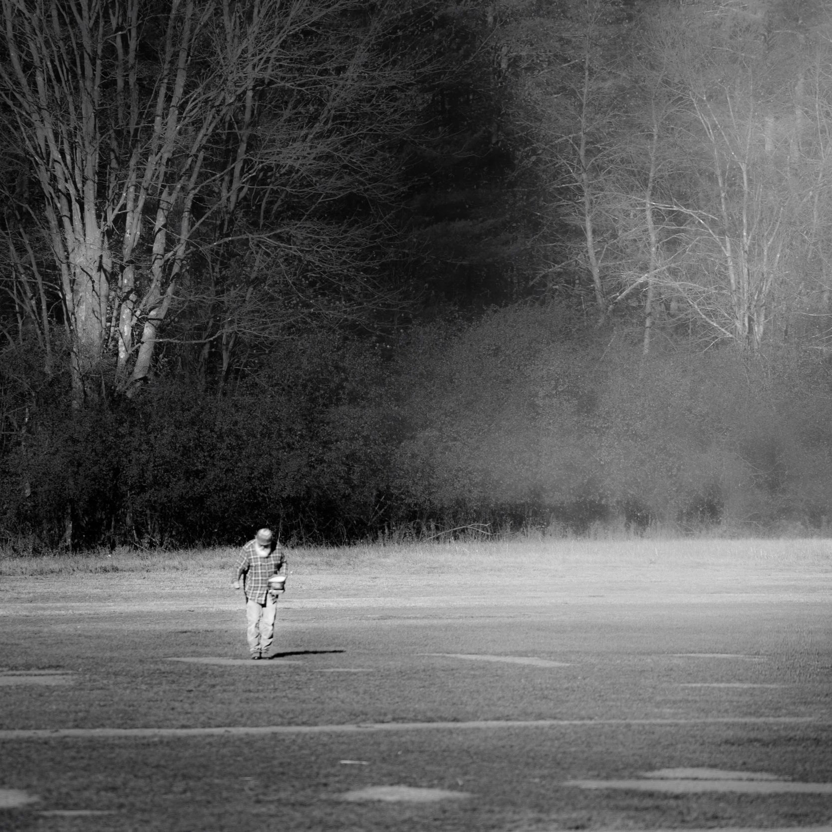 A person in a plaid shirt and a hat walking across a large open field with trees in the background