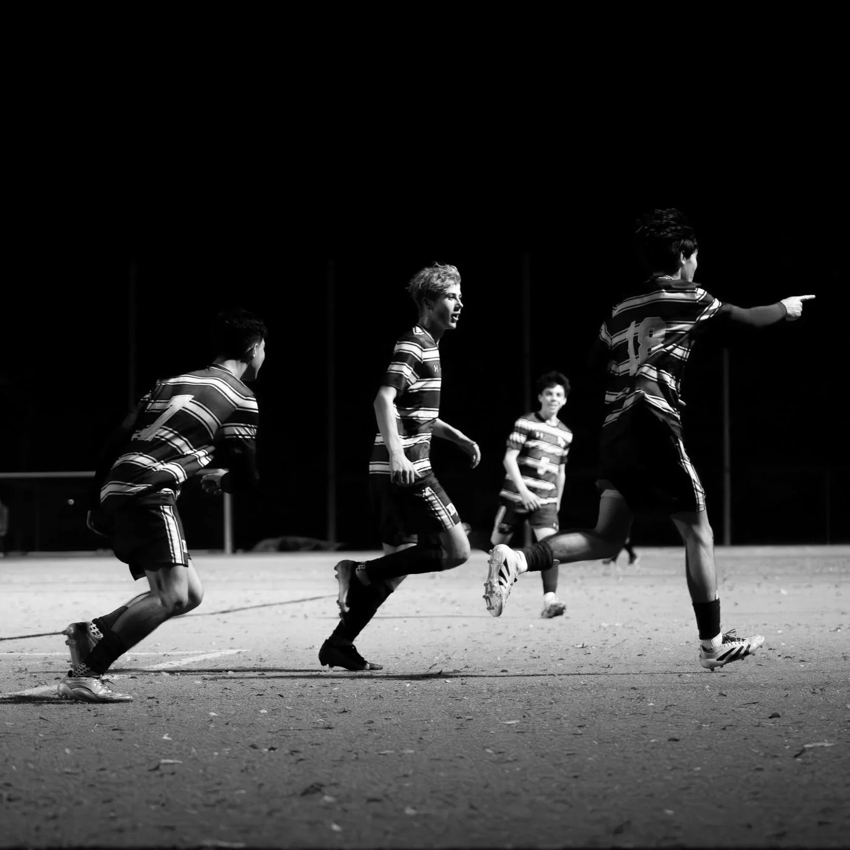 Black and white photo of children playing soccer on a field at night, with one child captured mid-run pointing forward.