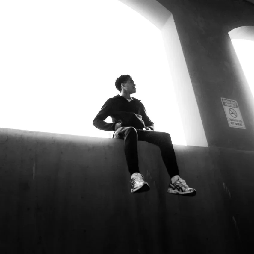 A young man sits on the edge of a concrete ledge in an outdoor urban setting, looking to the side with a contemplative expression. The photo is in black and white, with a large archway and a sign visible in the background.