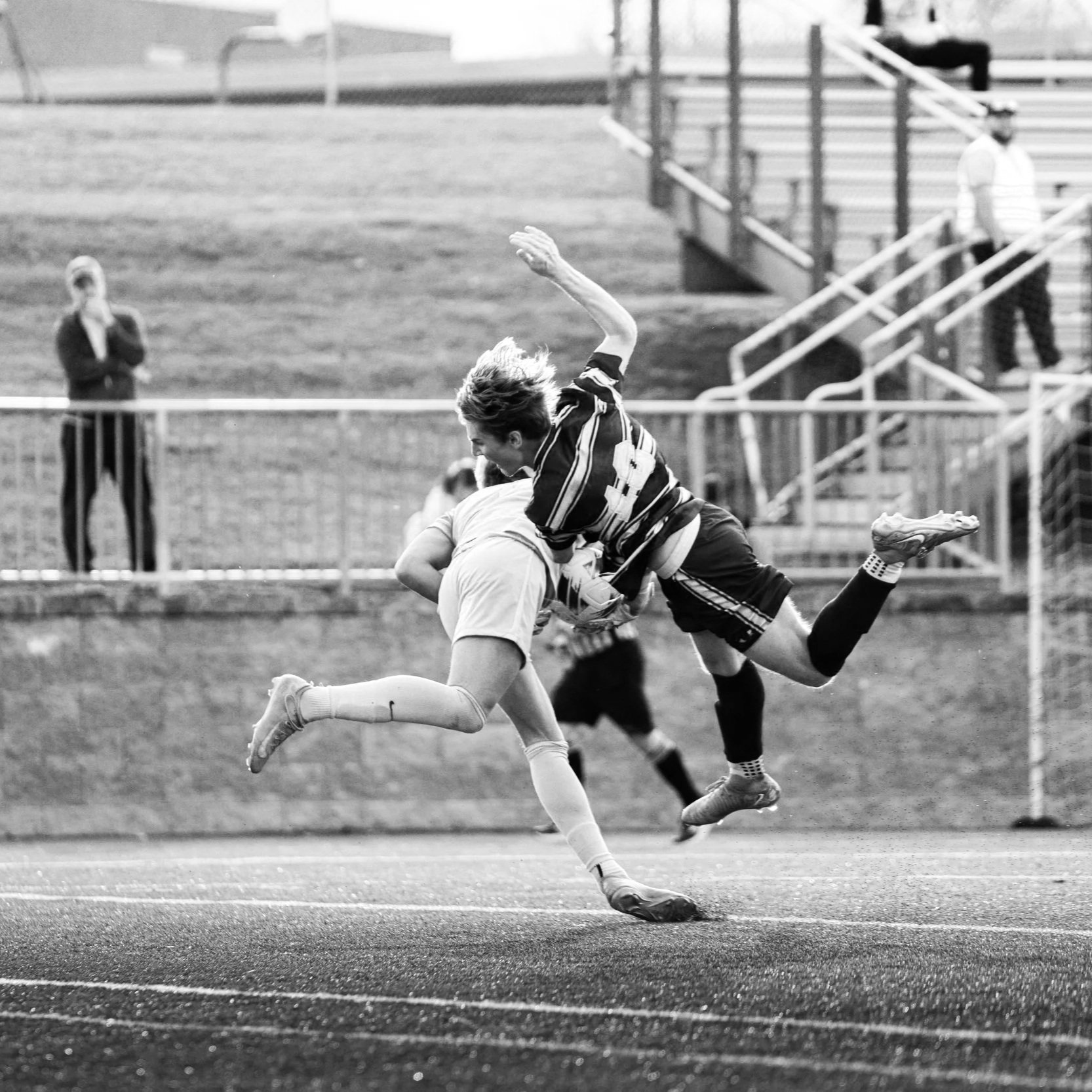 A black and white photo of two soccer players competing for the ball on an outdoor field. One player is mid-air, while the other is crouched down. Spectators are seen in the background near the bleachers.
