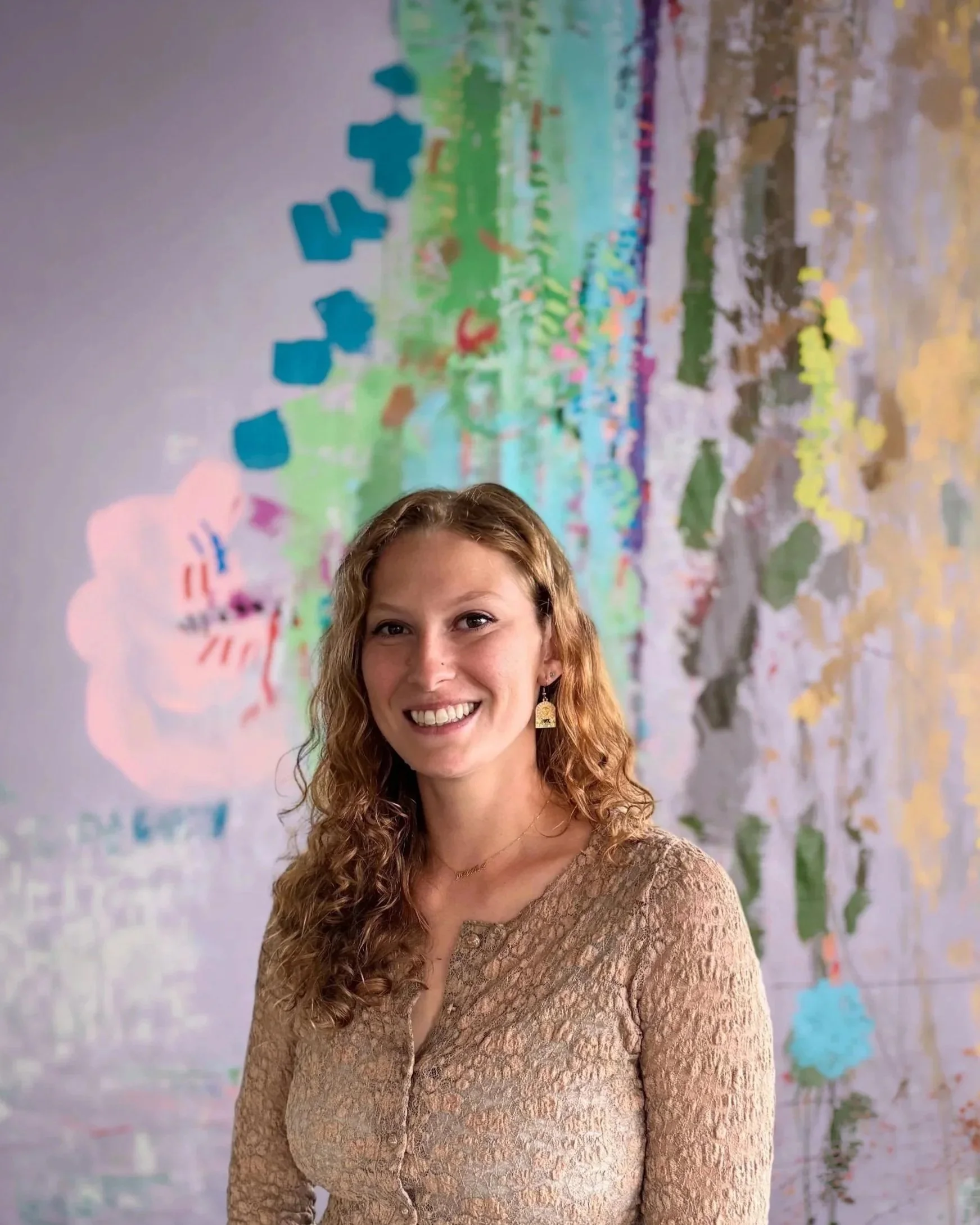 A woman with long curly red hair smiling at the camera, wearing a beige textured lace dress, standing in front of a colorful mural painting.