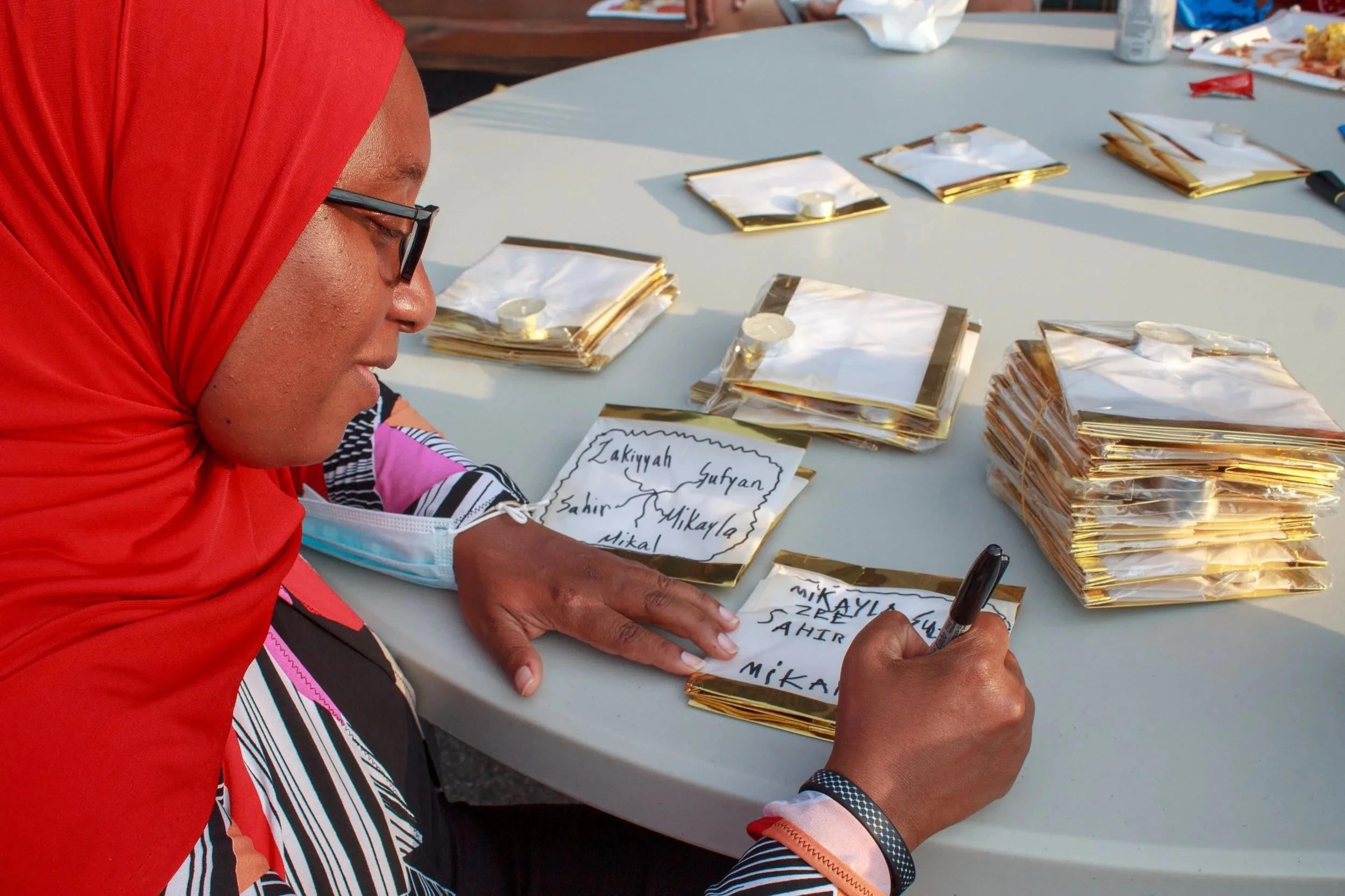 A woman with glasses and a red headscarf writes names on small gold-colored framed papers at a table. Multiple similar framed papers and a marker are on the table.