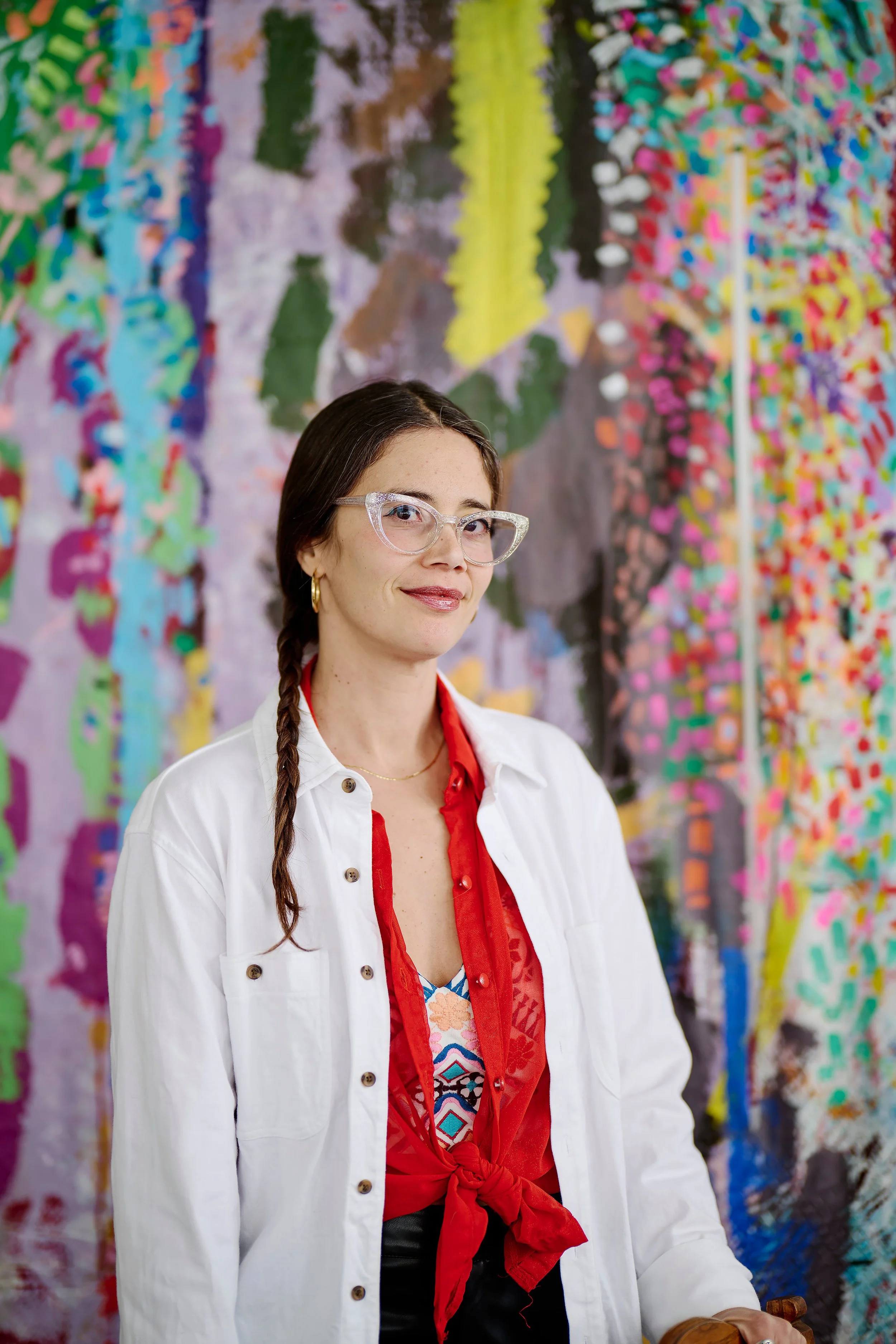 A woman with long dark hair, wearing glasses, a white jacket, and a red shirt, stands in front of a colorful, abstract artwork.