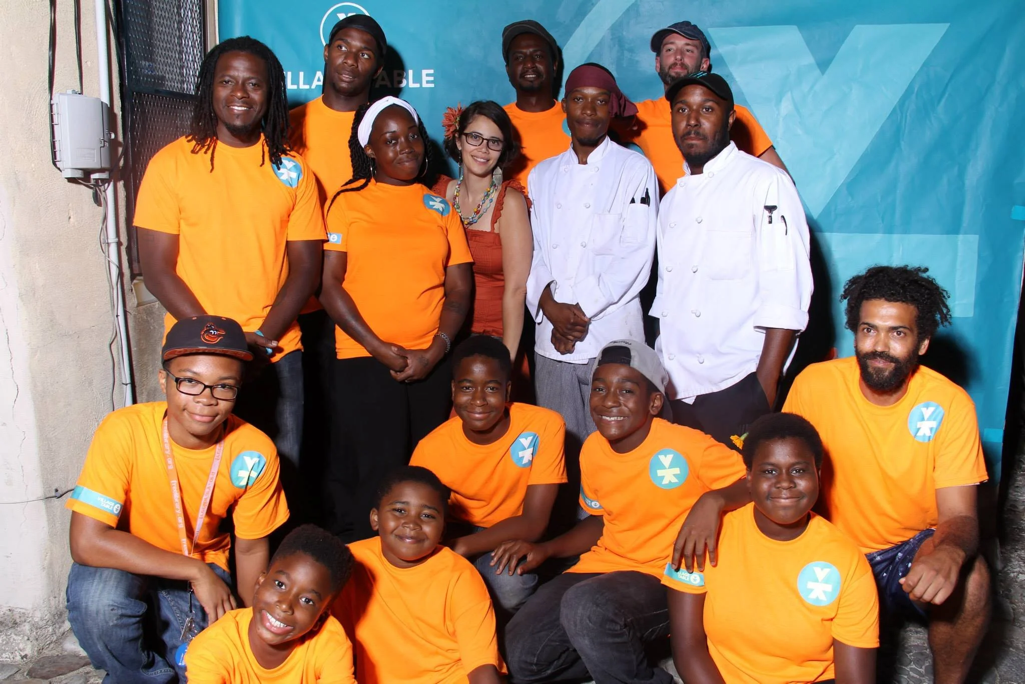 Group of diverse young people, some in orange shirts and others in white chef uniforms, posing together at an event against a blue backdrop. Many are smiling, and some wear hats or glasses.