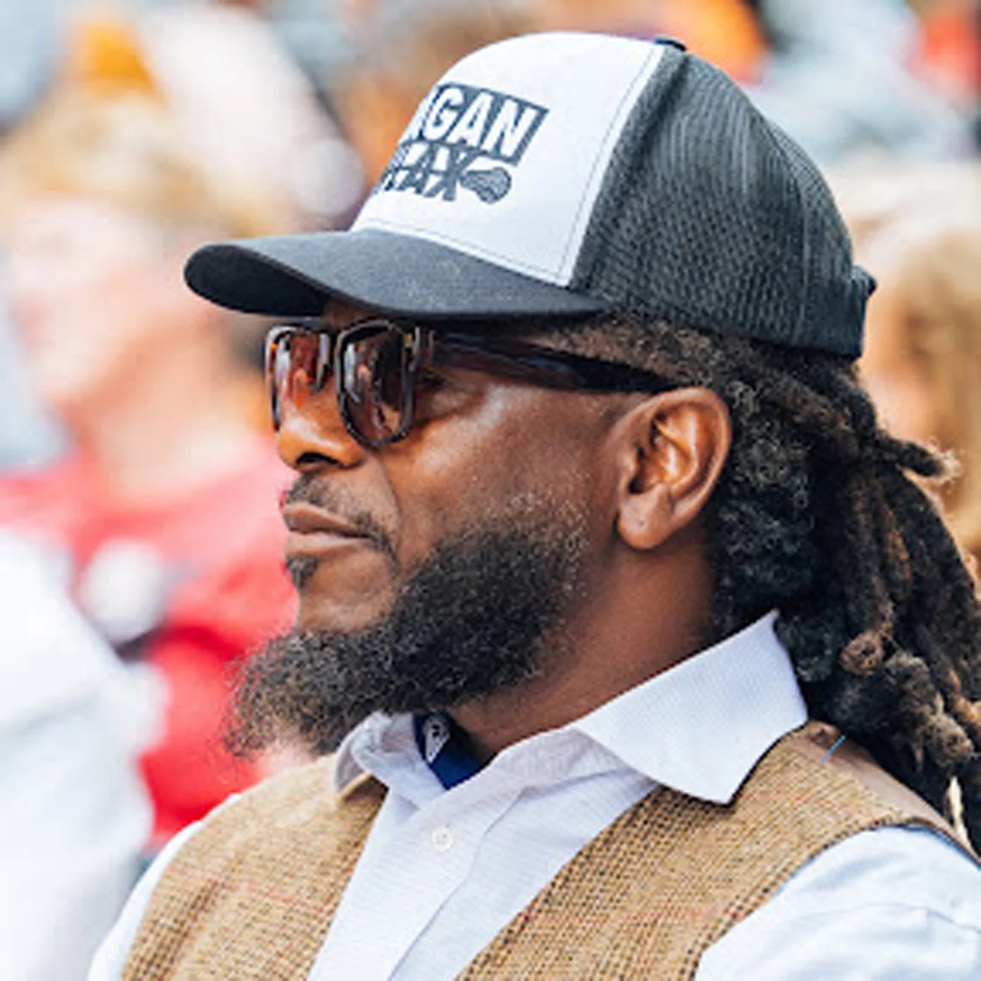 A man with dreadlocks wearing sunglasses, a trucker hat, a white shirt, and a brown vest in profile view.