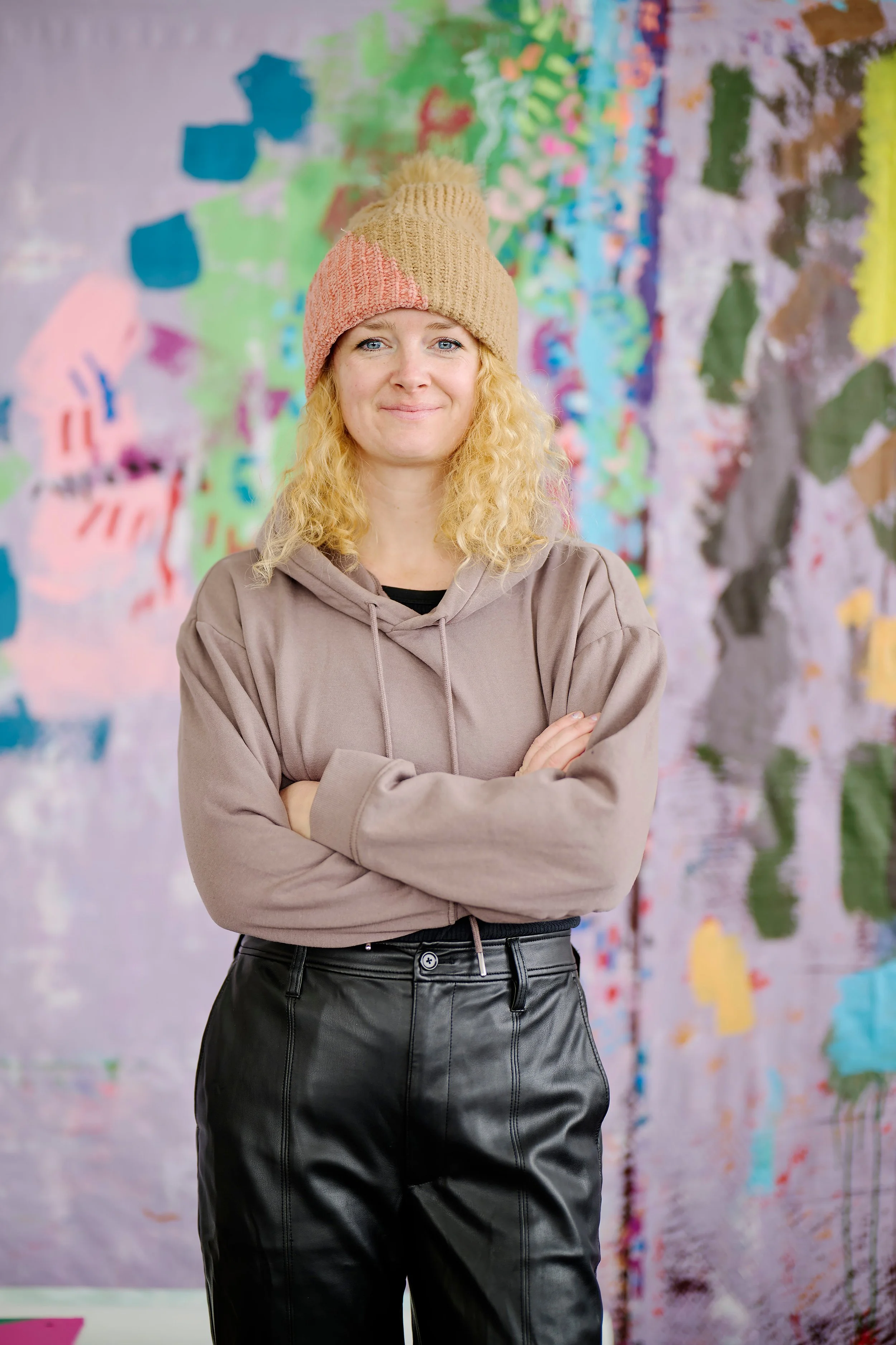 A woman with blonde curly hair wearing a brown knit beanie, a beige hoodie, and black leather pants stands with her arms crossed in front of a colorful abstract art background.