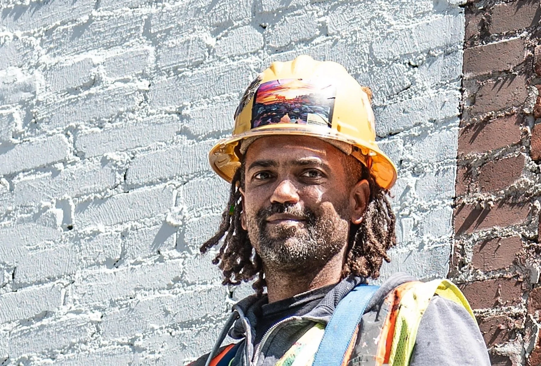 A construction worker with dreadlocks and a beard taking a selfie in front of a textured wall with a mix of painted and brick sections. He is wearing a yellow safety helmet and a reflective safety vest.
