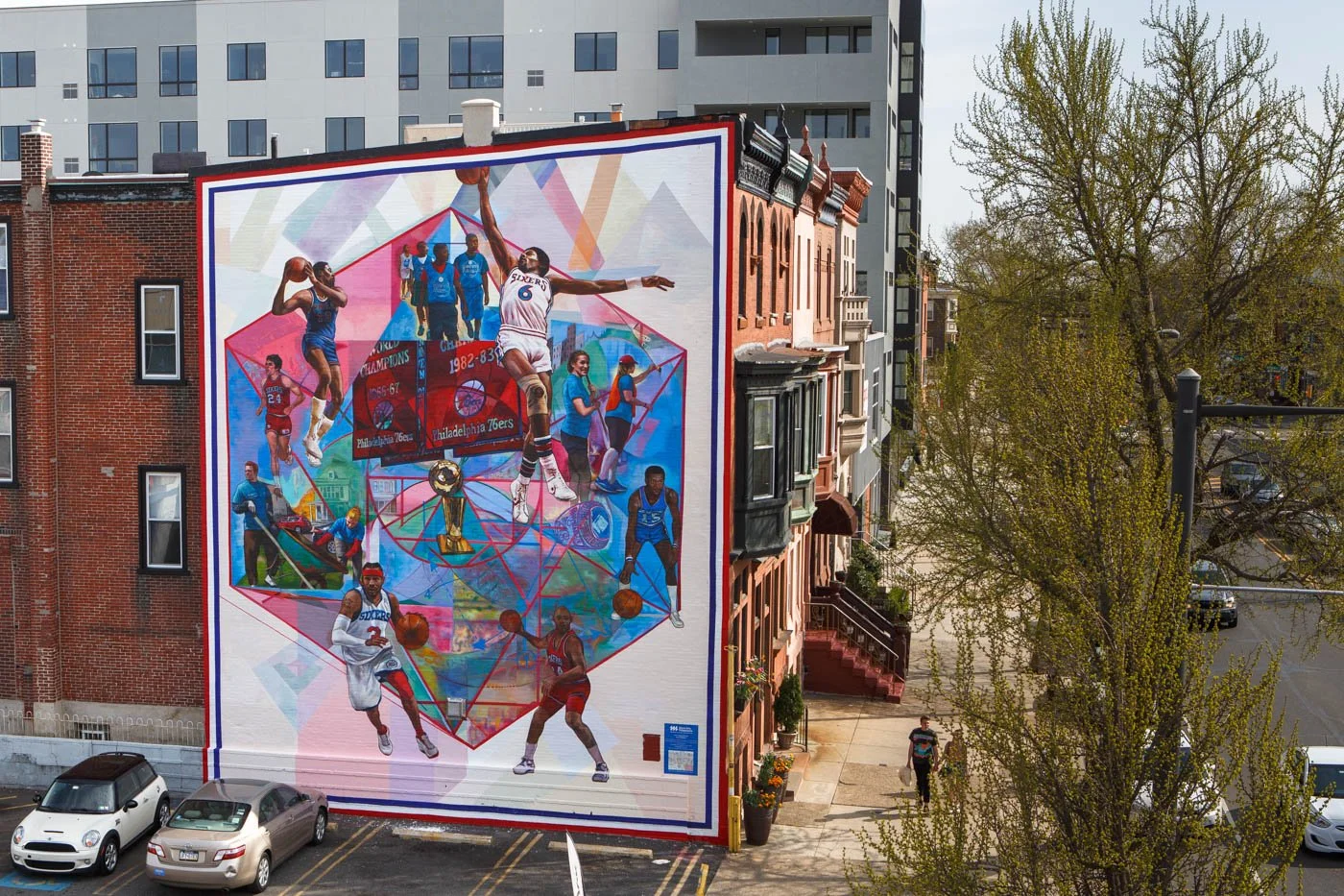 Colorful mural celebrating the Philadelphia 76ers basketball team featuring players in action, a trophy, and historic team moments painted on side of a building in an urban neighborhood.