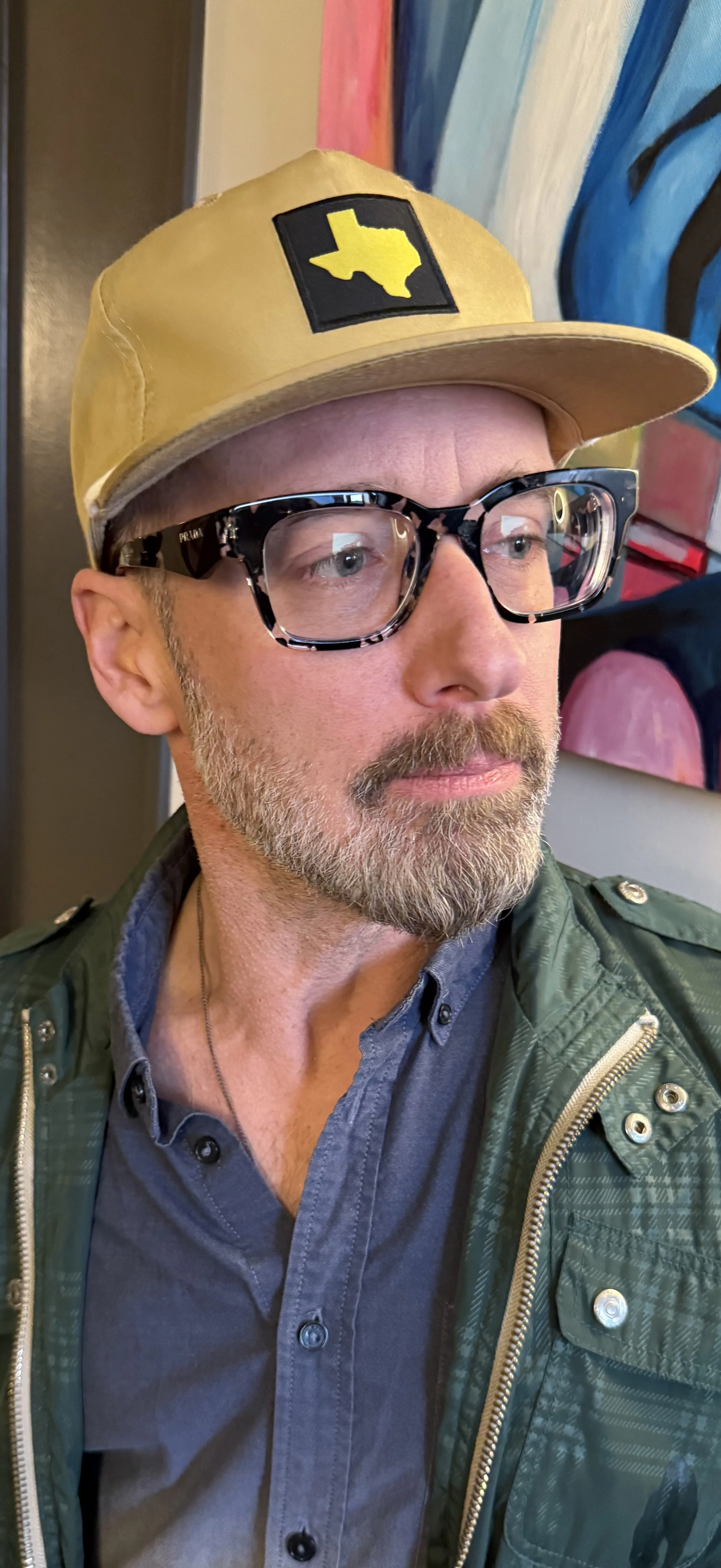 A man with glasses, a beard, and mustache wearing a yellow cap with a Texas-shaped patch, a gray button-up shirt, and a green jacket. He is indoors with abstract artwork in the background.