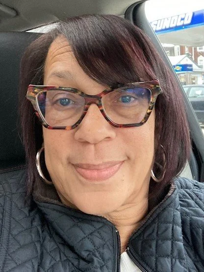 A woman with glasses and hoop earrings smiling inside a car, with a Sunoco gas station visible through the window behind her.