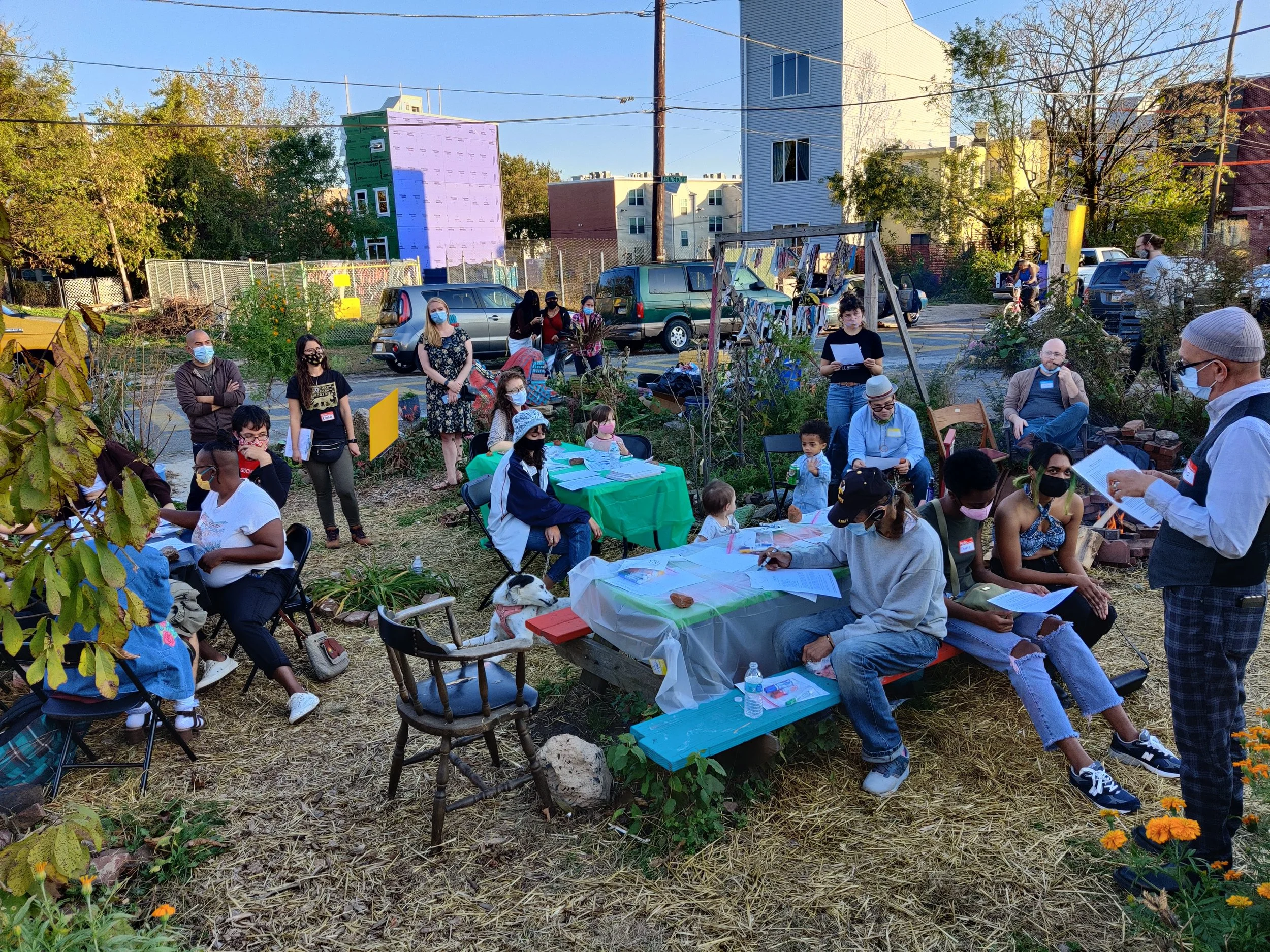 A community gathering outdoors during the daytime, with a group of people wearing masks, some sitting at tables and others standing, in an urban garden setting with colorful mural building and residential structures in the background.