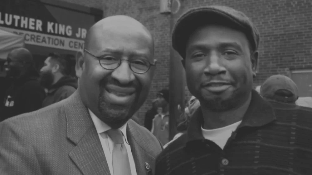 Two men smiling at a gathering outside a building with a sign that reads "Luther King Jr. Recreation Center." One man is wearing glasses, a suit, and a tie; the other man is wearing a hat and a striped shirt.
