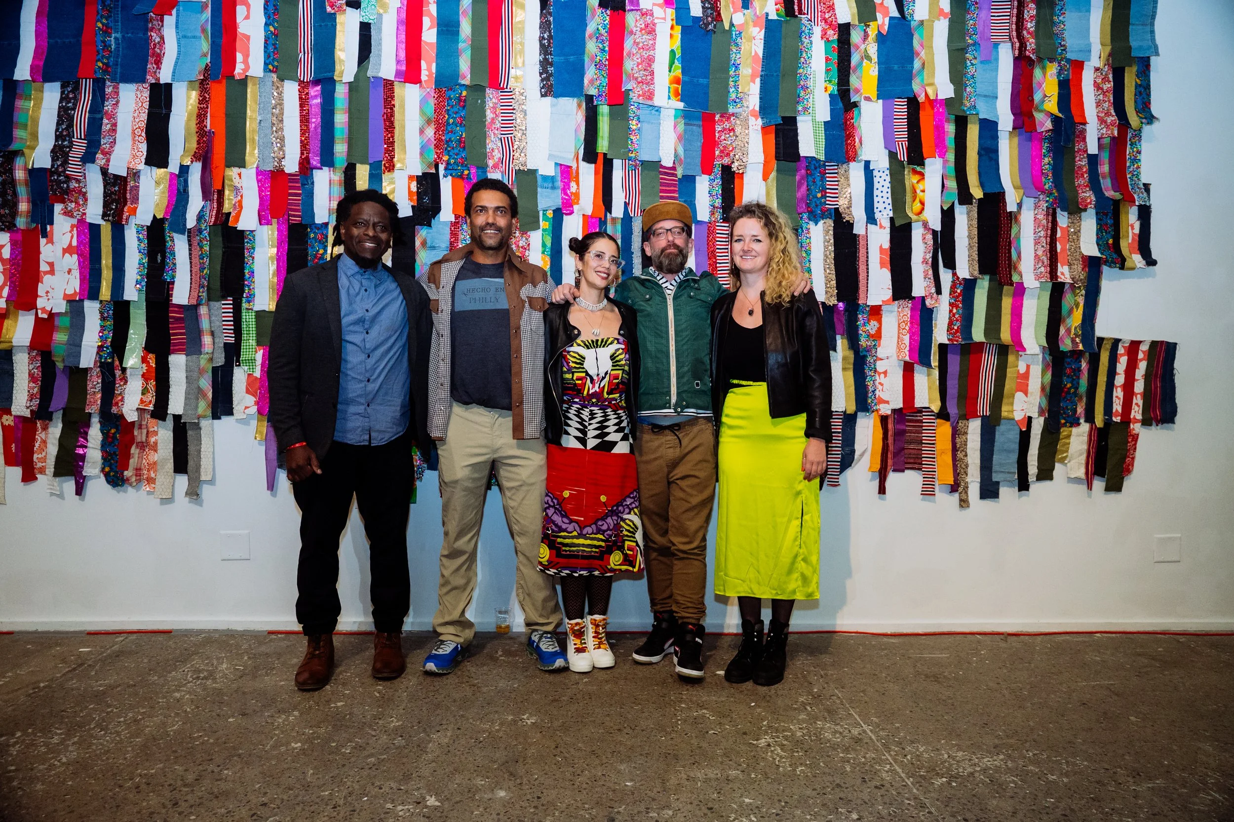 Five people standing in front of a colorful wall hanging made of various patterned fabric strips. They are smiling and dressed in a mix of casual and eclectic clothing.