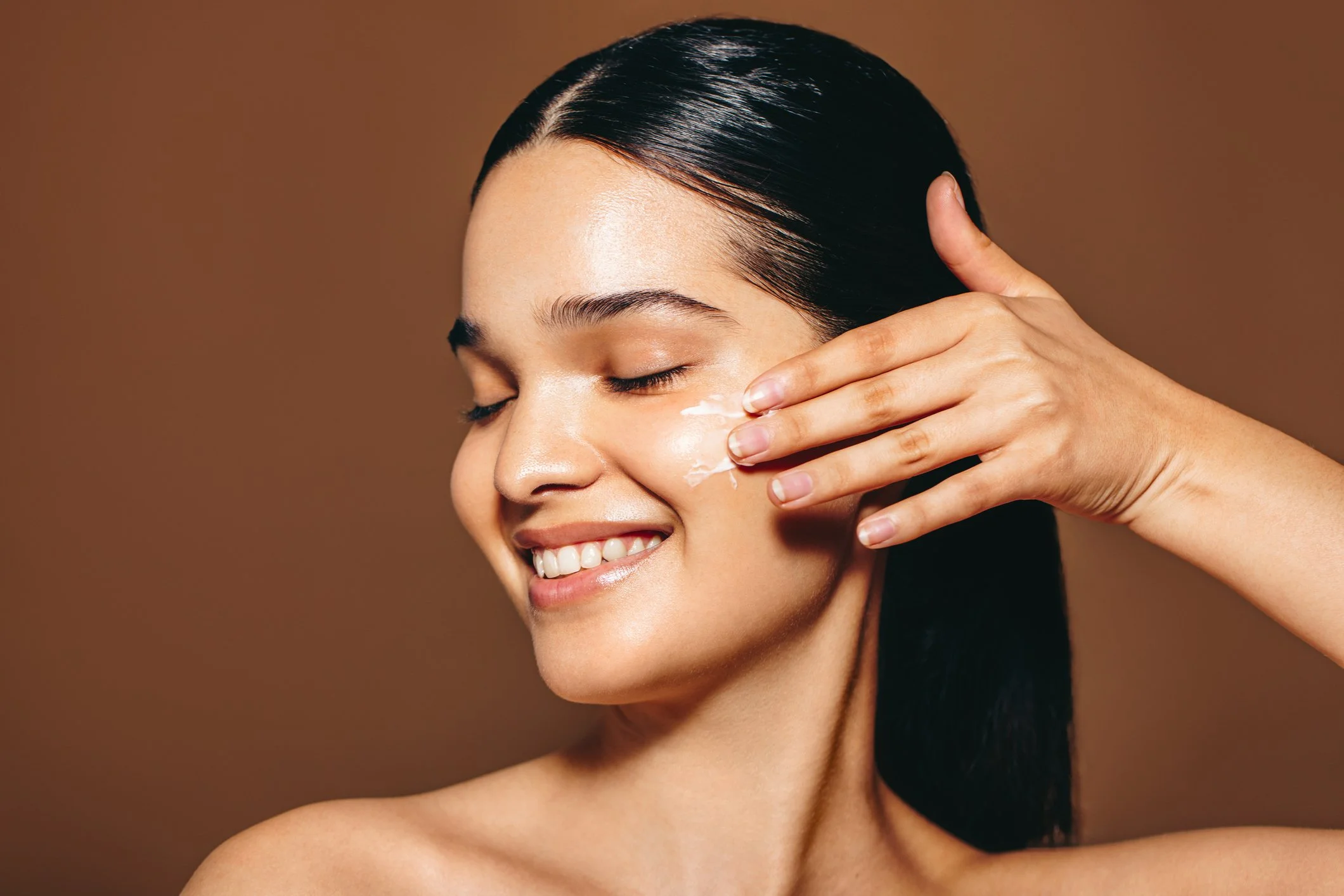A smiling woman with closed eyes and dark, wet hair, gently touching her face with her fingers on a brown background.