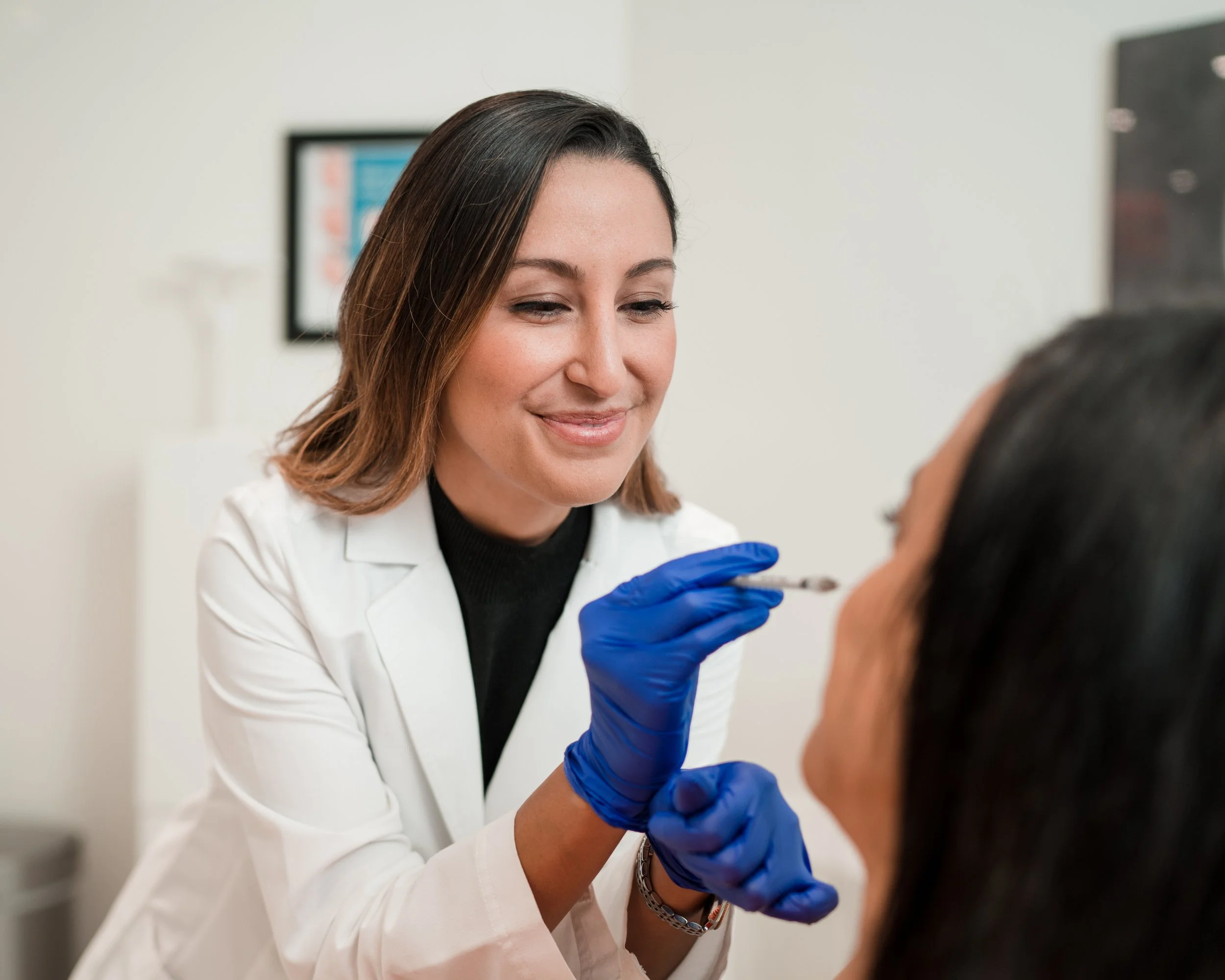 A female healthcare professional wearing a white lab coat and blue gloves prepares to give a vaccination injection to a patient. The woman has shoulder-length brown hair and is smiling gently. The background is an indoor clinical setting with a framed chart or poster on the wall.