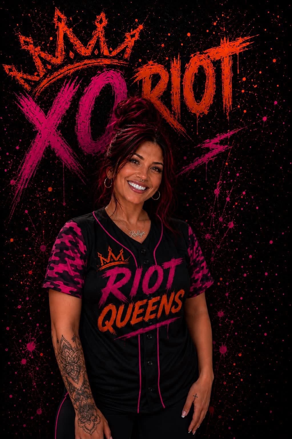 A woman standing in front of a black background with pink and orange graffiti-style text and graphics reading 'Riot Queens.' She is smiling, has a nose ring, hoop earrings, and tattoos on her arm and hand. She is wearing a black and pink jersey-style shirt with the same text and graphics, and a necklace that spells 'Rilyn.'