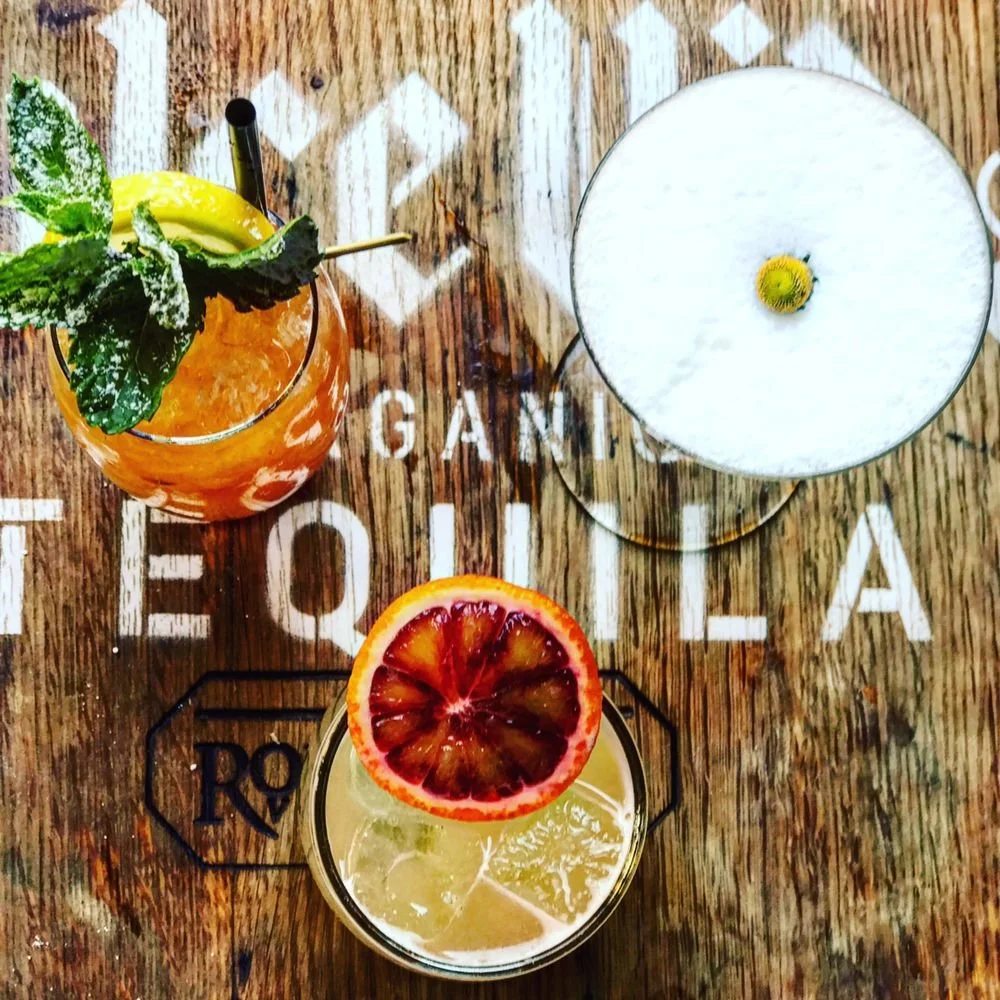 Top view of a wooden table with three colorful cocktails. One with mint and lemon garnish, one with blood orange slice, and one with a lemon slice and frothy foam on top.