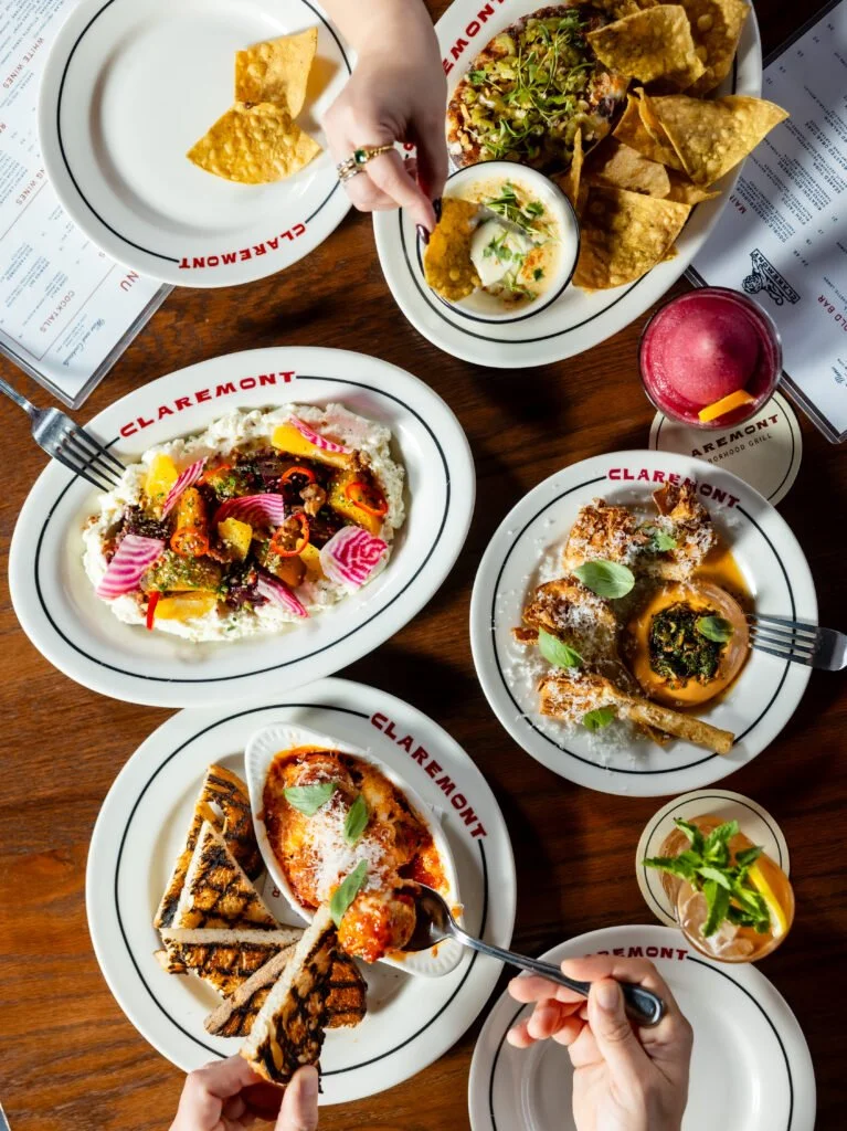 A variety of dishes including nachos with cheese, a salad with colorful vegetables, fried chicken with sauce, grilled flatbread, a pink drink with orange peel, and a small glass of a cocktail with mint. The dishes are served on white plates with the restaurant's name "Claremont".