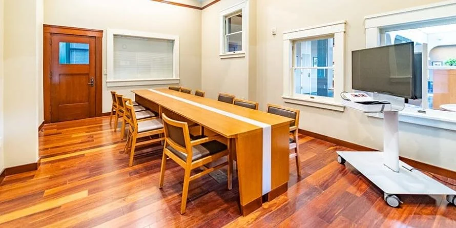 Conference room with a long wooden table, several chairs, a flat-screen monitor on a mobile stand, and windows.