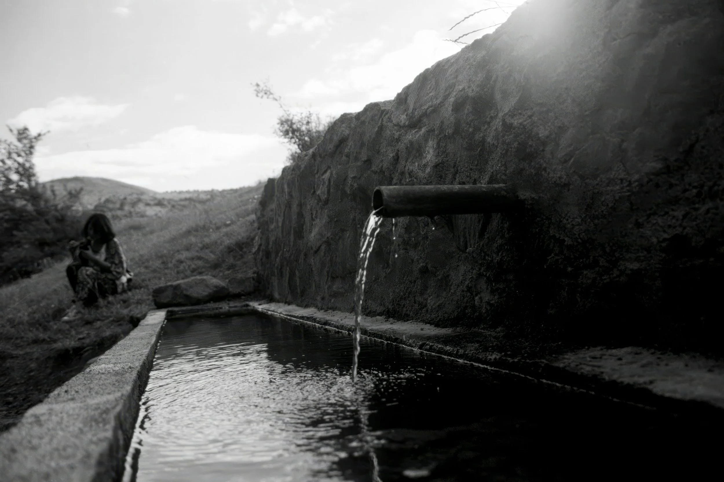 A metal pipe protrudes from a rocky hillside, releasing water into a concrete flood drain below. A person is sitting on the ground to the left of the drain, with natural landscape and hills in the background.