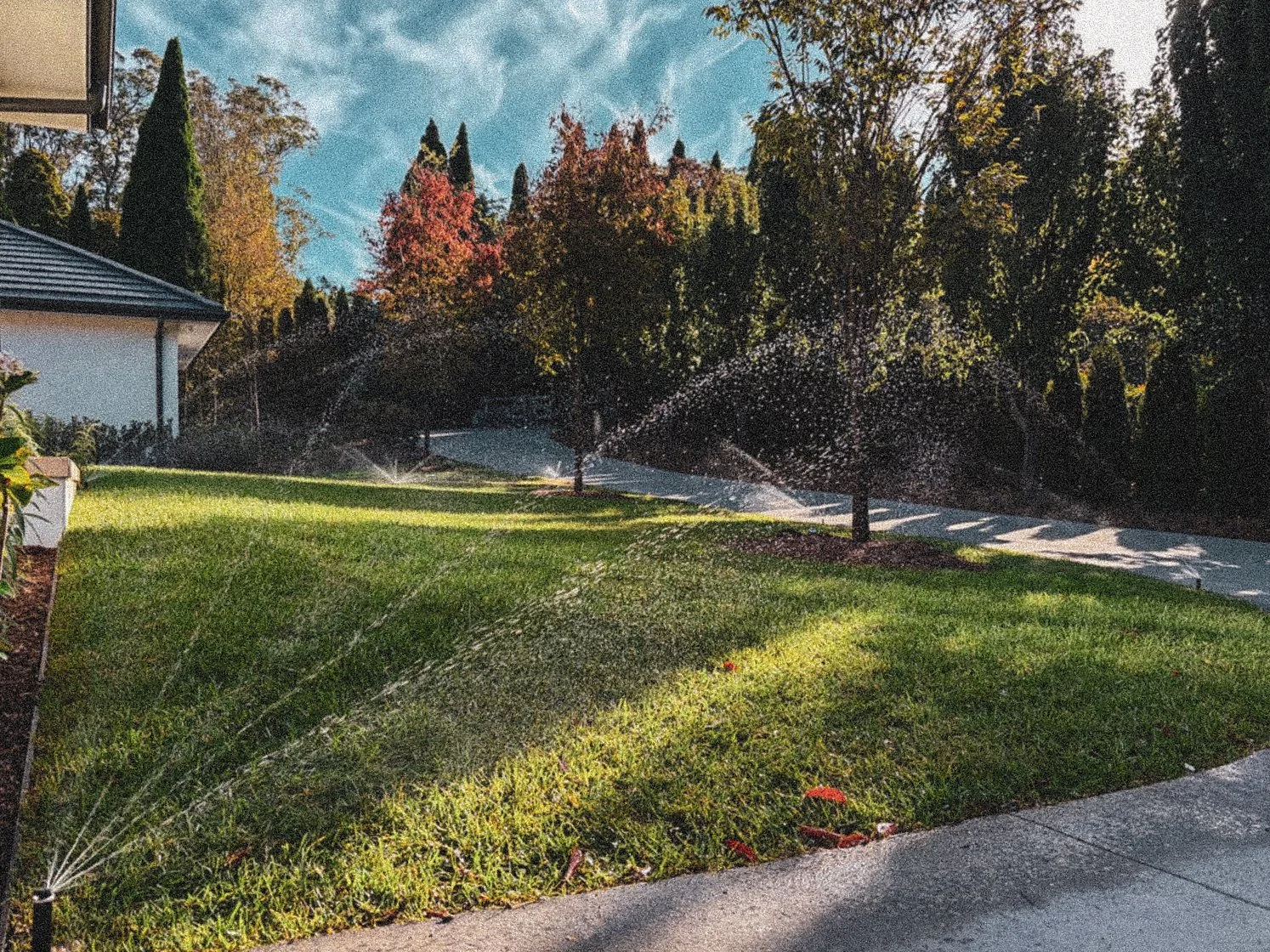 A backyard with a well-kept green lawn, several sprinklers watering the grass, and trees with fall foliage. A sidewalk runs through the yard, and part of a house with a roof is visible on the left. The sky is partly cloudy with blue skies and white clouds.
