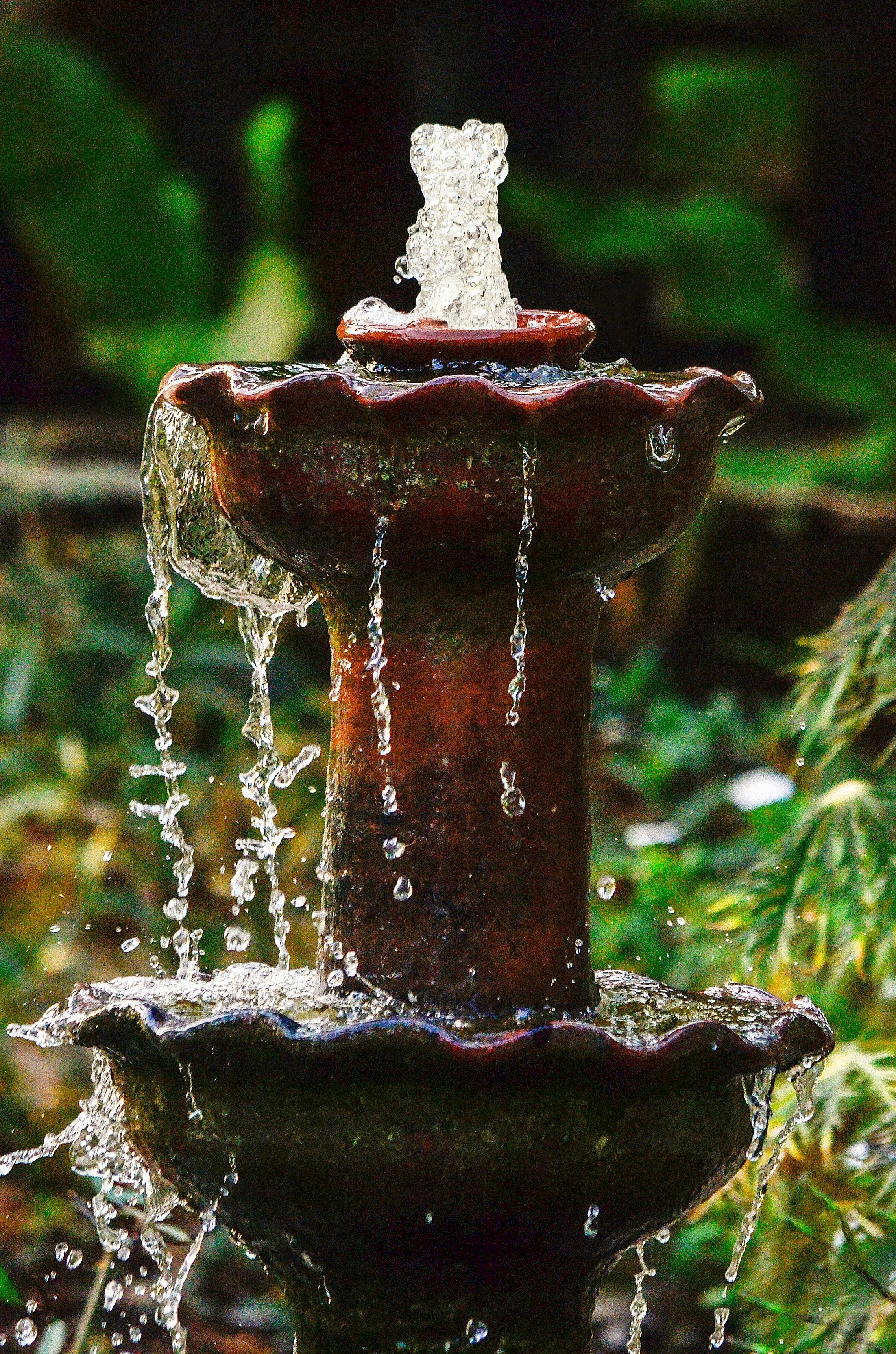 Close-up of a tiered outdoor water fountain with water cascading down the dark ceramic tiers, set against a background of green foliage.