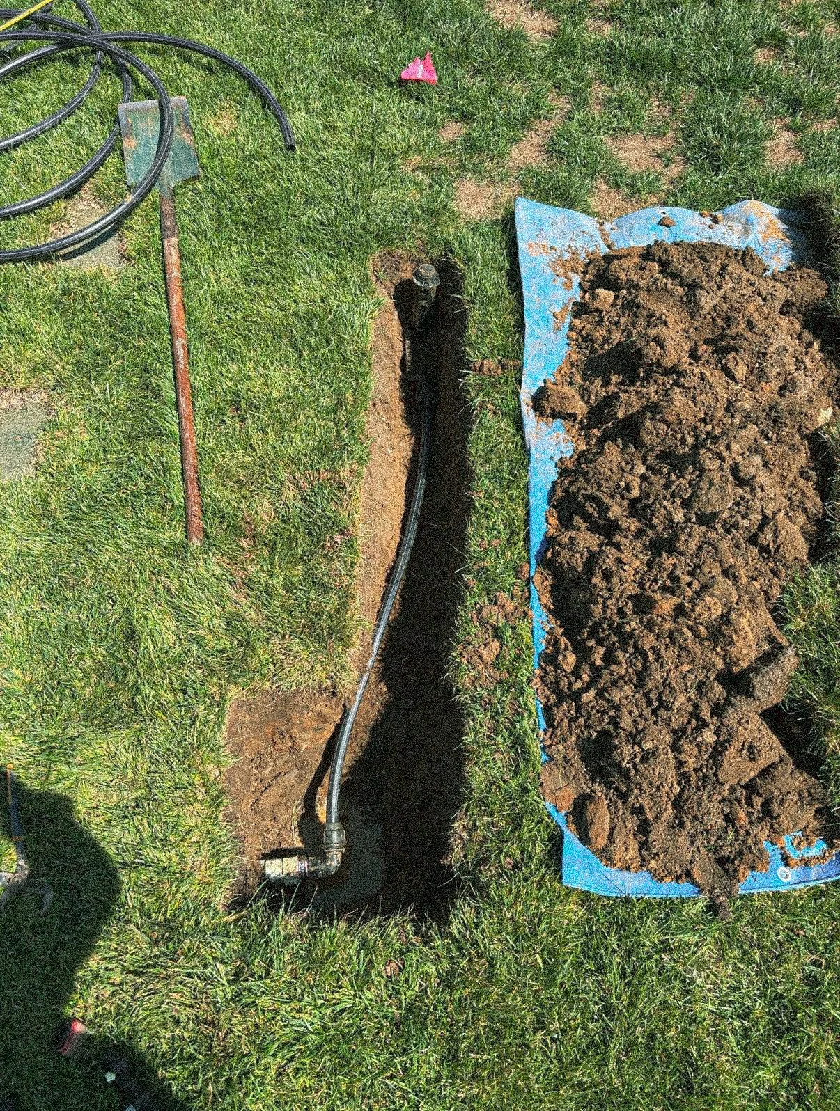 A backyard with a trench and a black pipeline being installed underground. There's a pile of soil on a blue tarp and some garden tools and hoses nearby.