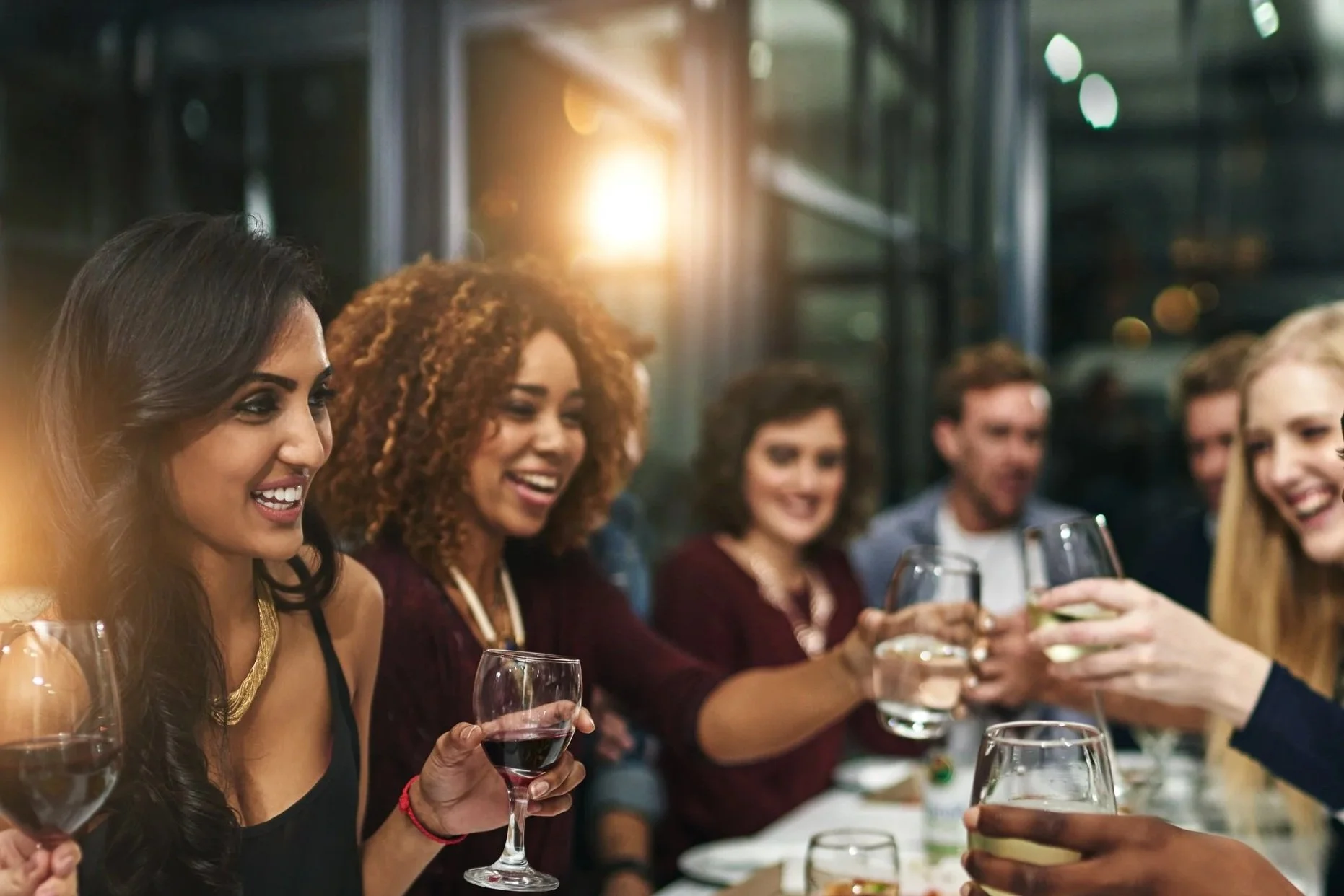 Group of diverse people at dinner party clinking wine glasses and smiling at each other in a restaurant with warm lighting.