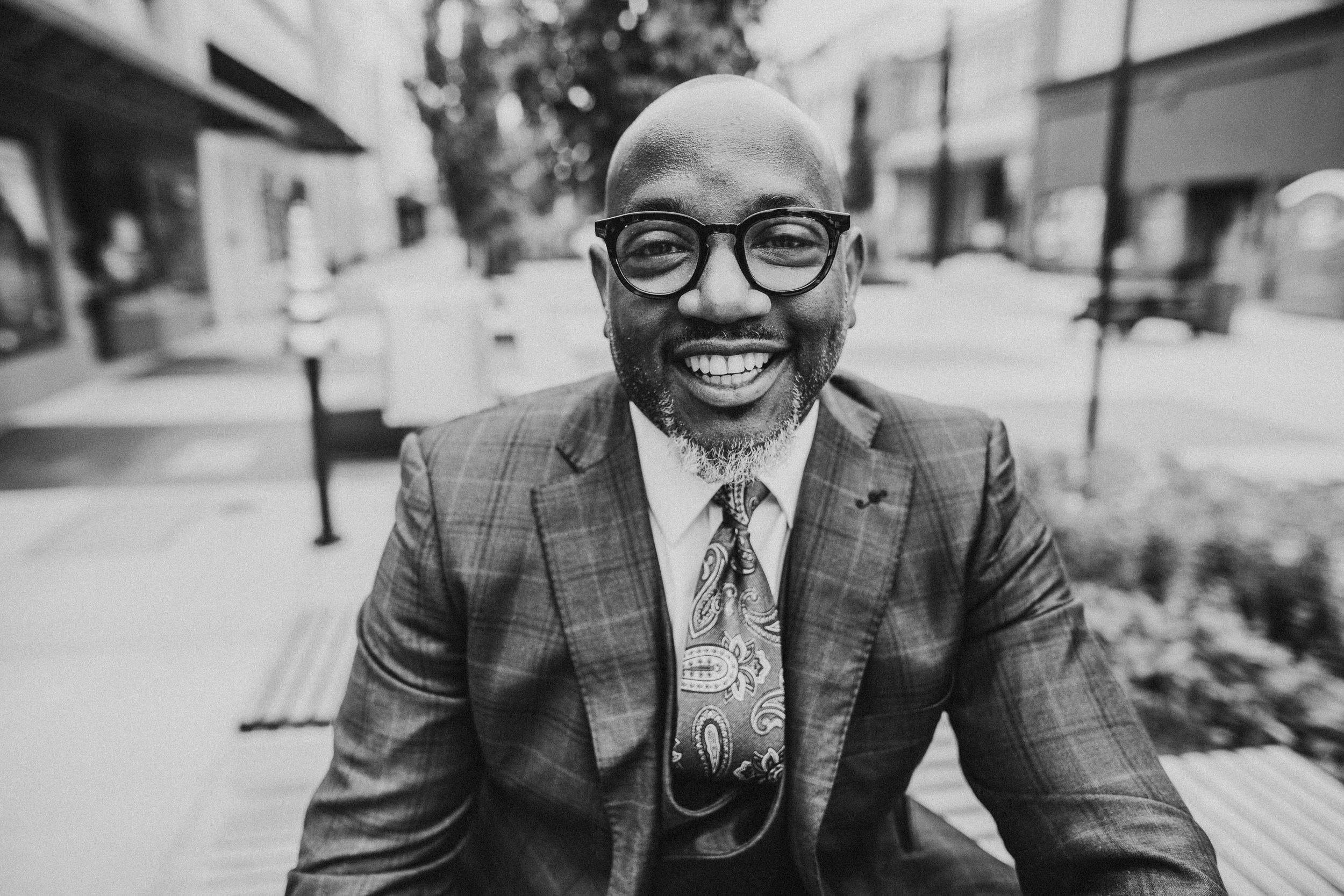 A smiling successful black man with glasses, dressed in a plaid blazer and paisley tie, seated outdoors in an urban environment with buildings and trees in the background, depicted in black and white.