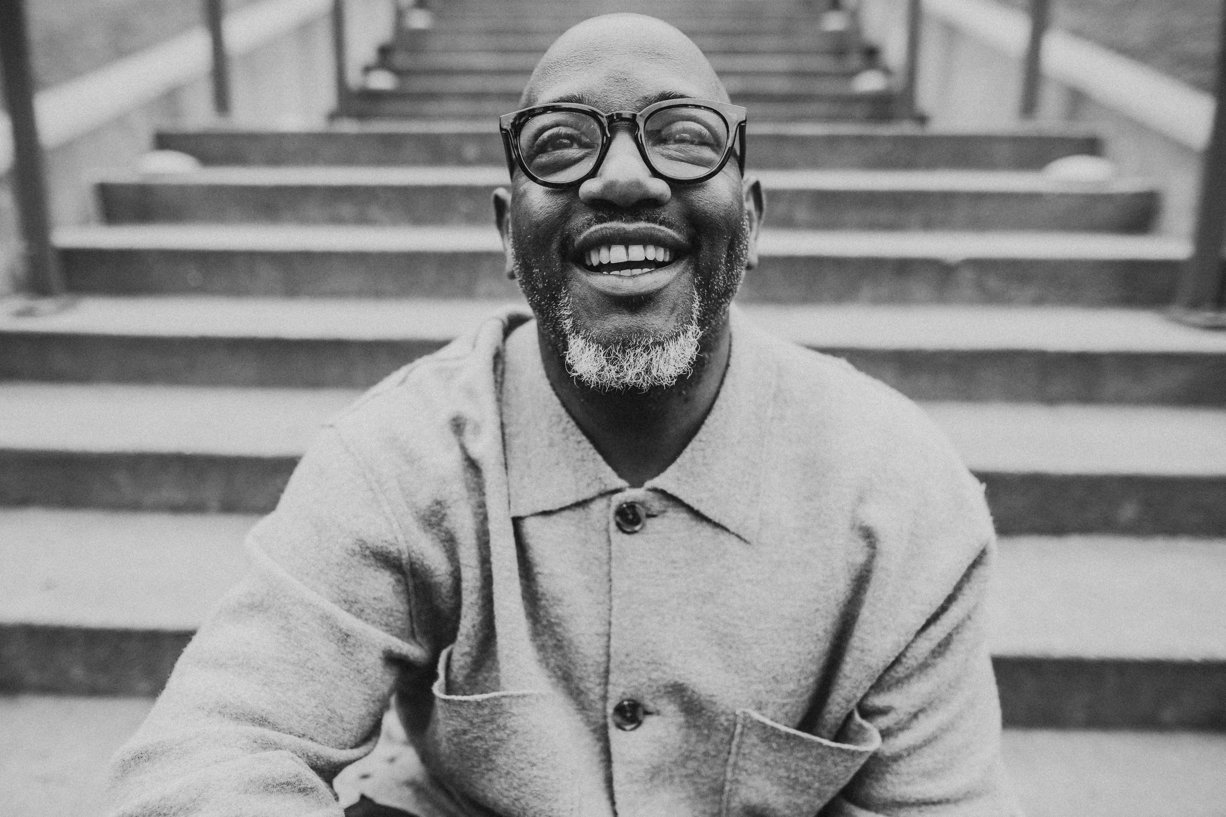 A close-up black and white photo of a smiling black man with glasses, sitting on outdoor stairs.