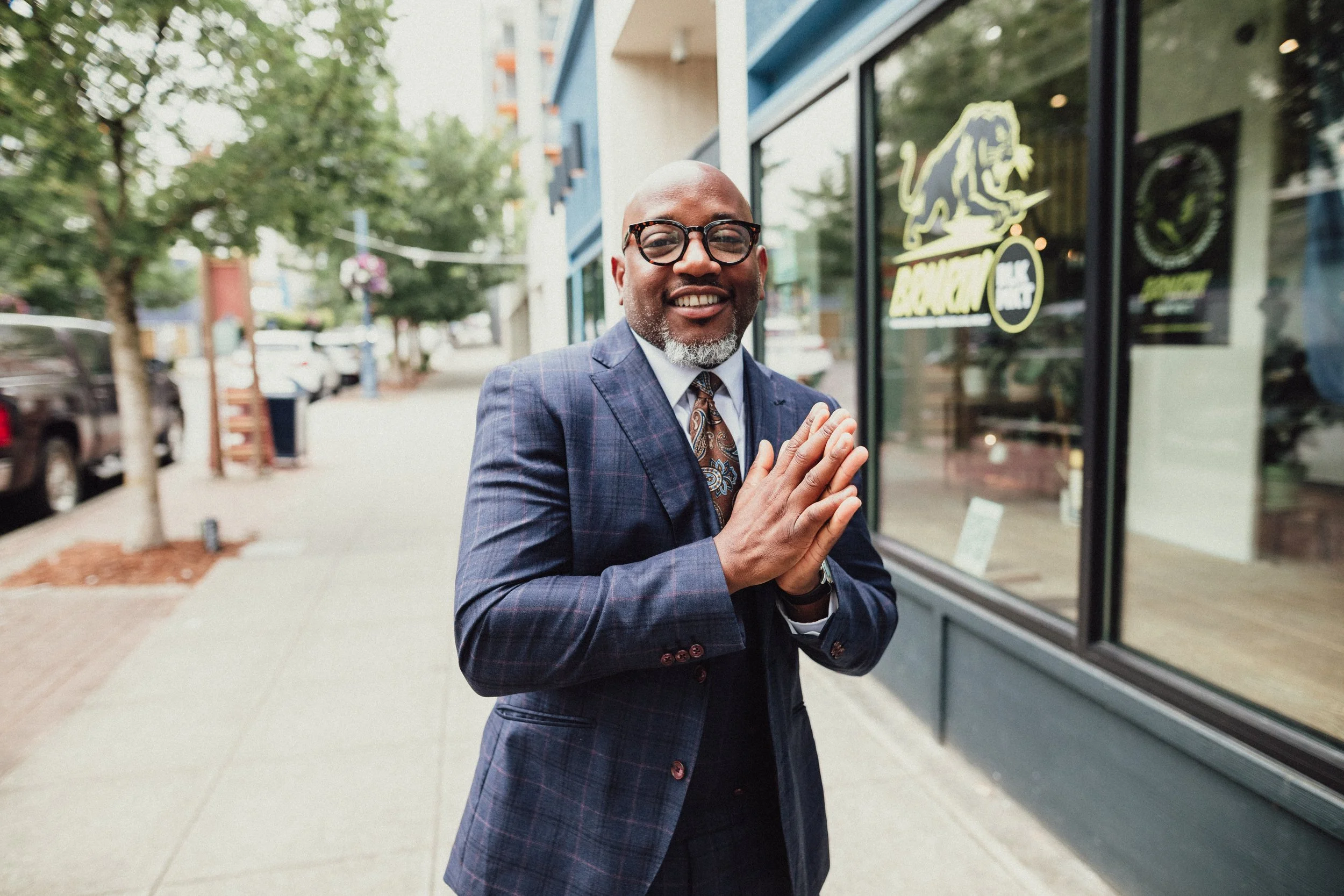 A smiling man with a bald head, glasses, and a gray beard, dressed in a blue plaid suit, standing outside a storefront with a neon sign of a lion and the text 'BLOXAM' on the window. The man has his hands pressed together in front of his chest in a greeting or prayer gesture.