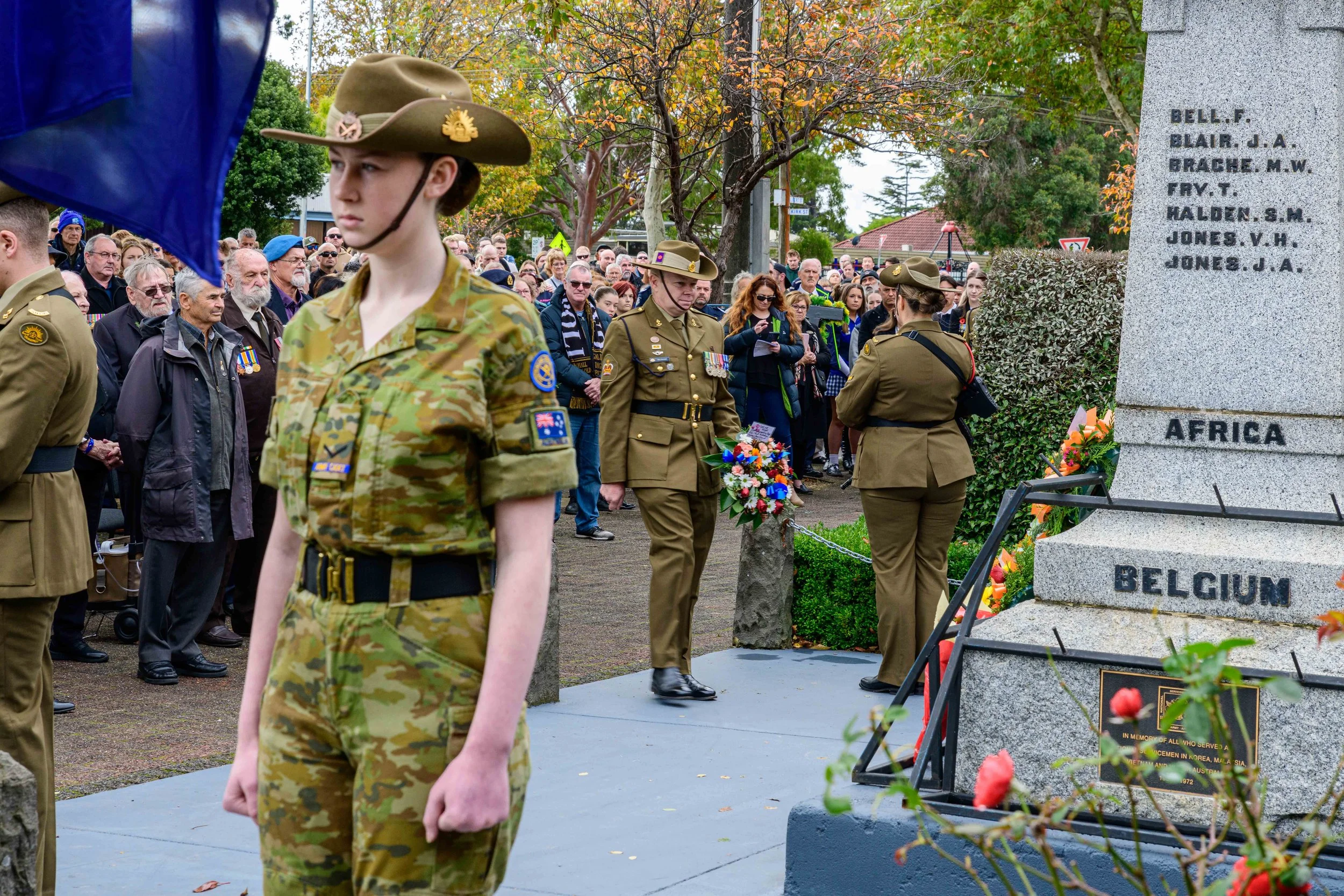 Military personnel paying respects at a memorial with a large crowd watching, trees with fall foliage in the background.