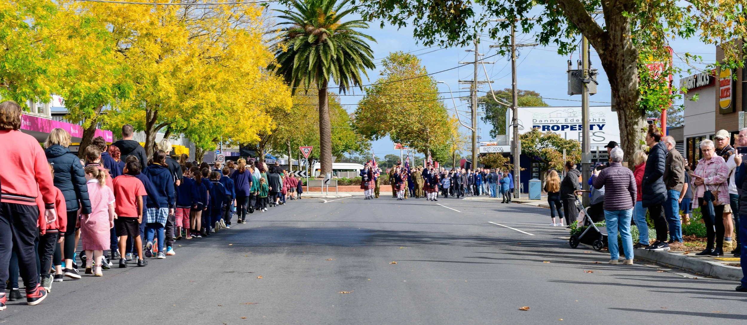 A street parade with children and adults watching as a bagpiper band marches in the background. The scene is set in autumn with yellow and green trees lining the street and people standing along the sidewalk.