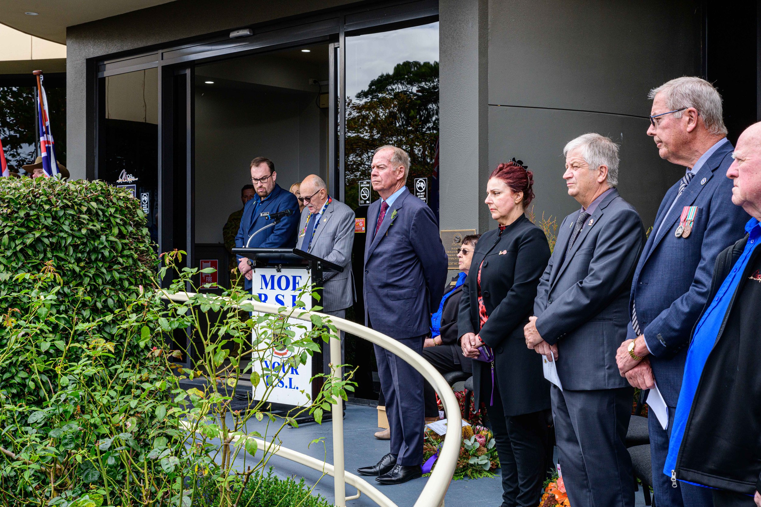 Group of people standing outdoors, paying respects during a memorial or remembrance event, with some wearing medals and formal attire, facing a speaker at a podium.